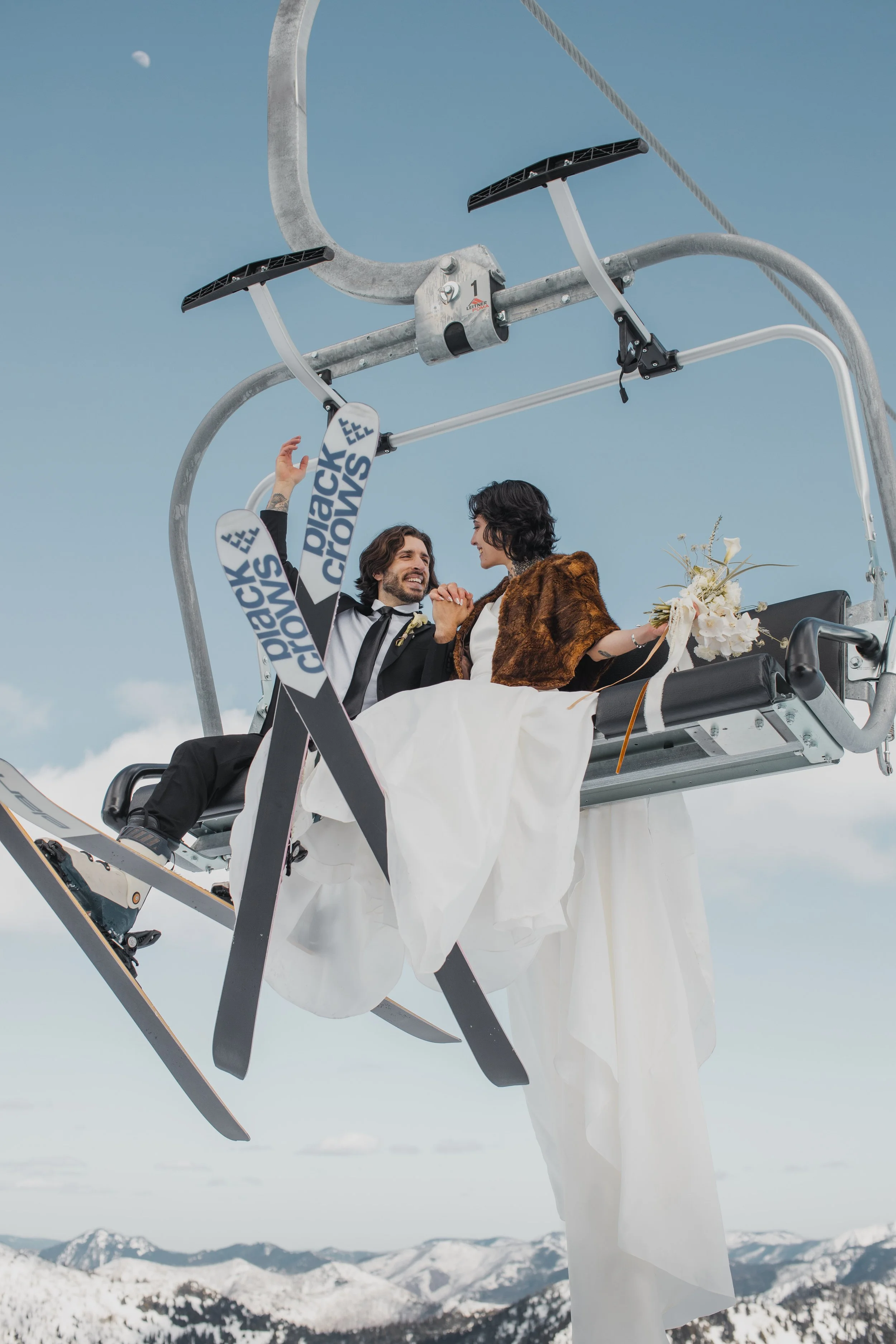 A couple in wedding attire riding a ski lift in a snowy mountain landscape with mountains in the background and a clear sky.
