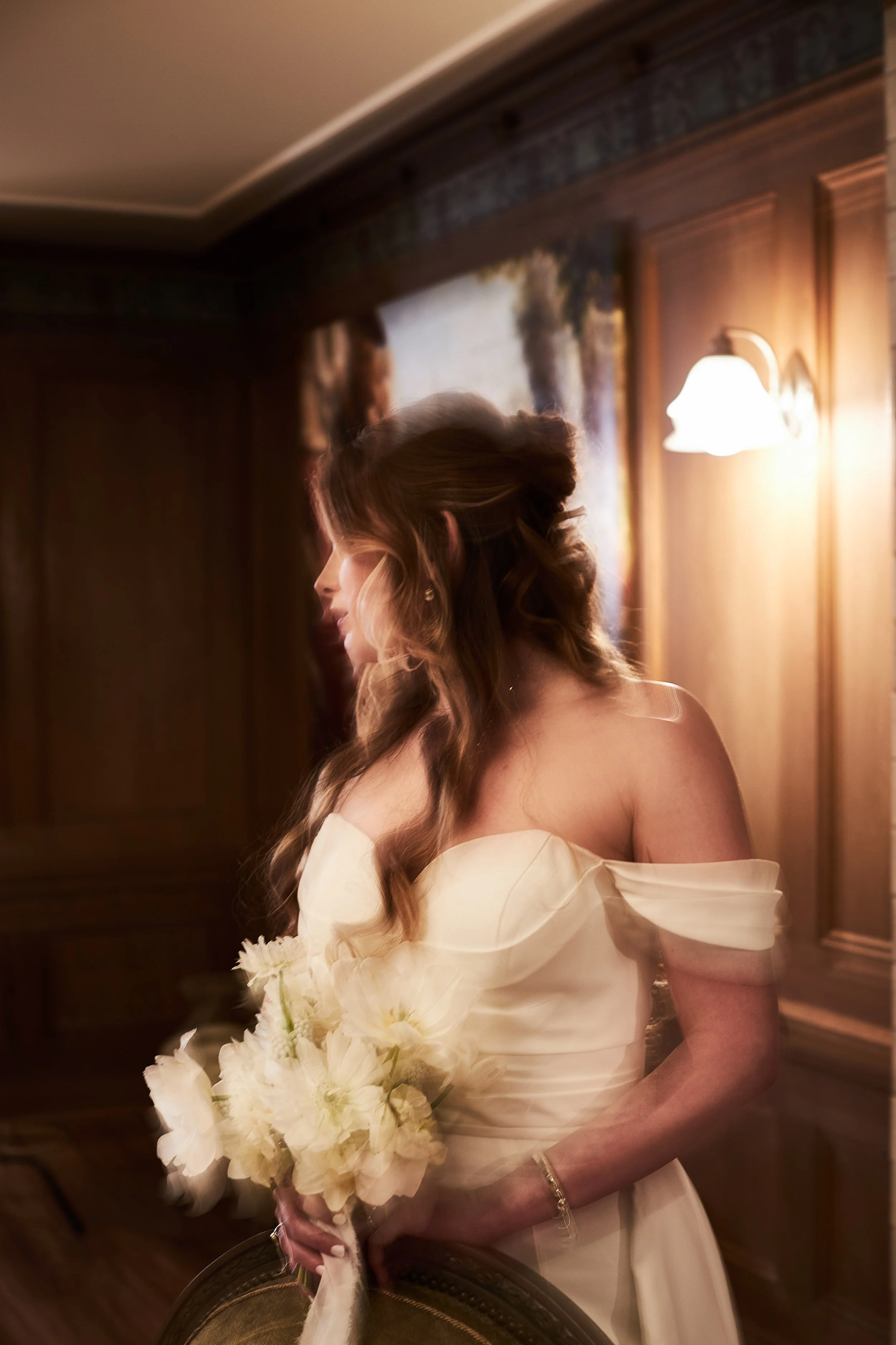 A woman in a wedding dress holding a bouquet of white flowers, standing in a warm-lit room with wooden walls and a wall-mounted lamp.