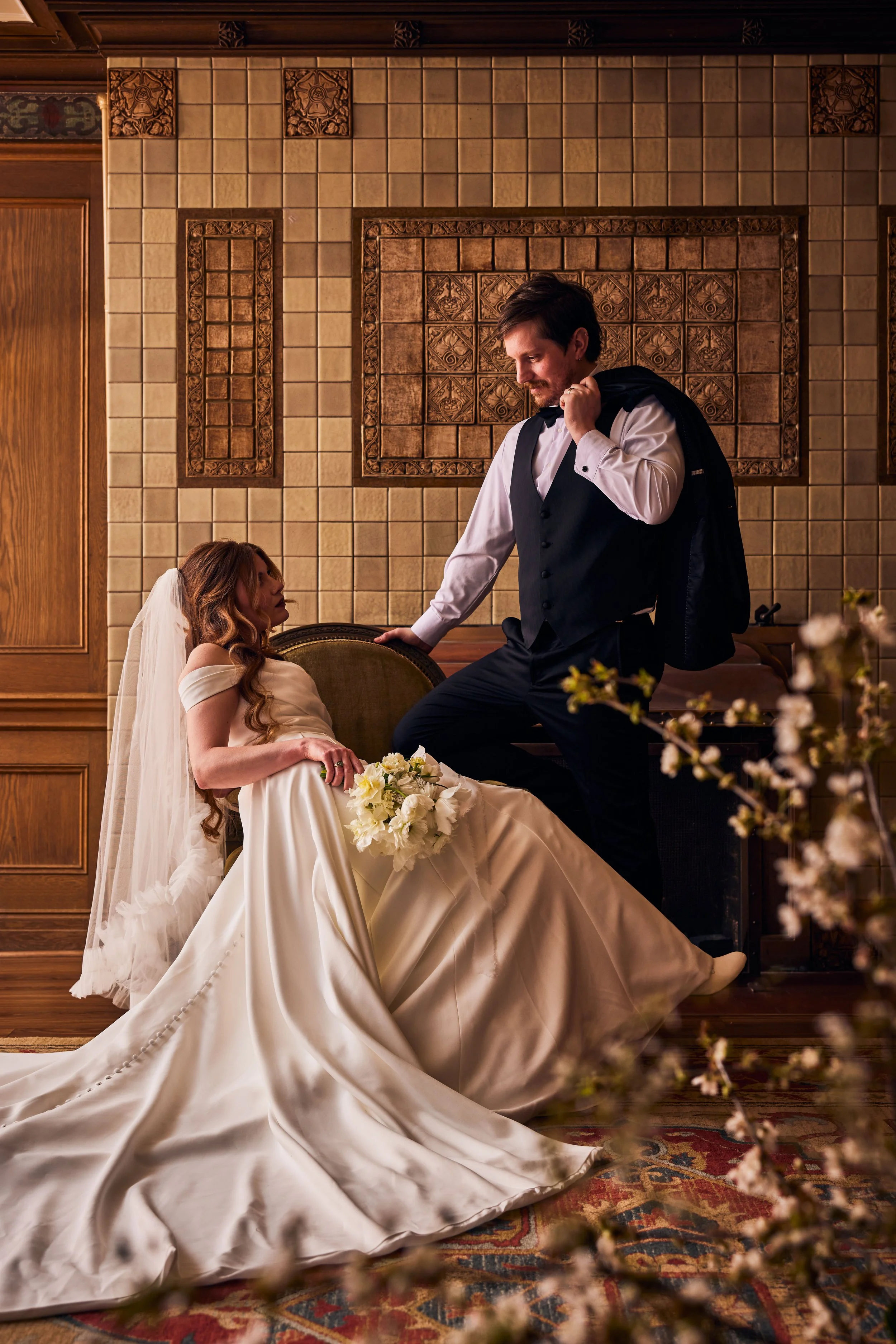 A bride sitting on a vintage-style chair with a white wedding gown, holding a bouquet of white flowers, while a man in a vest and dress shirt stands beside her, adjusting his jacket in an ornate, wood-paneled room with decorative tile wall.