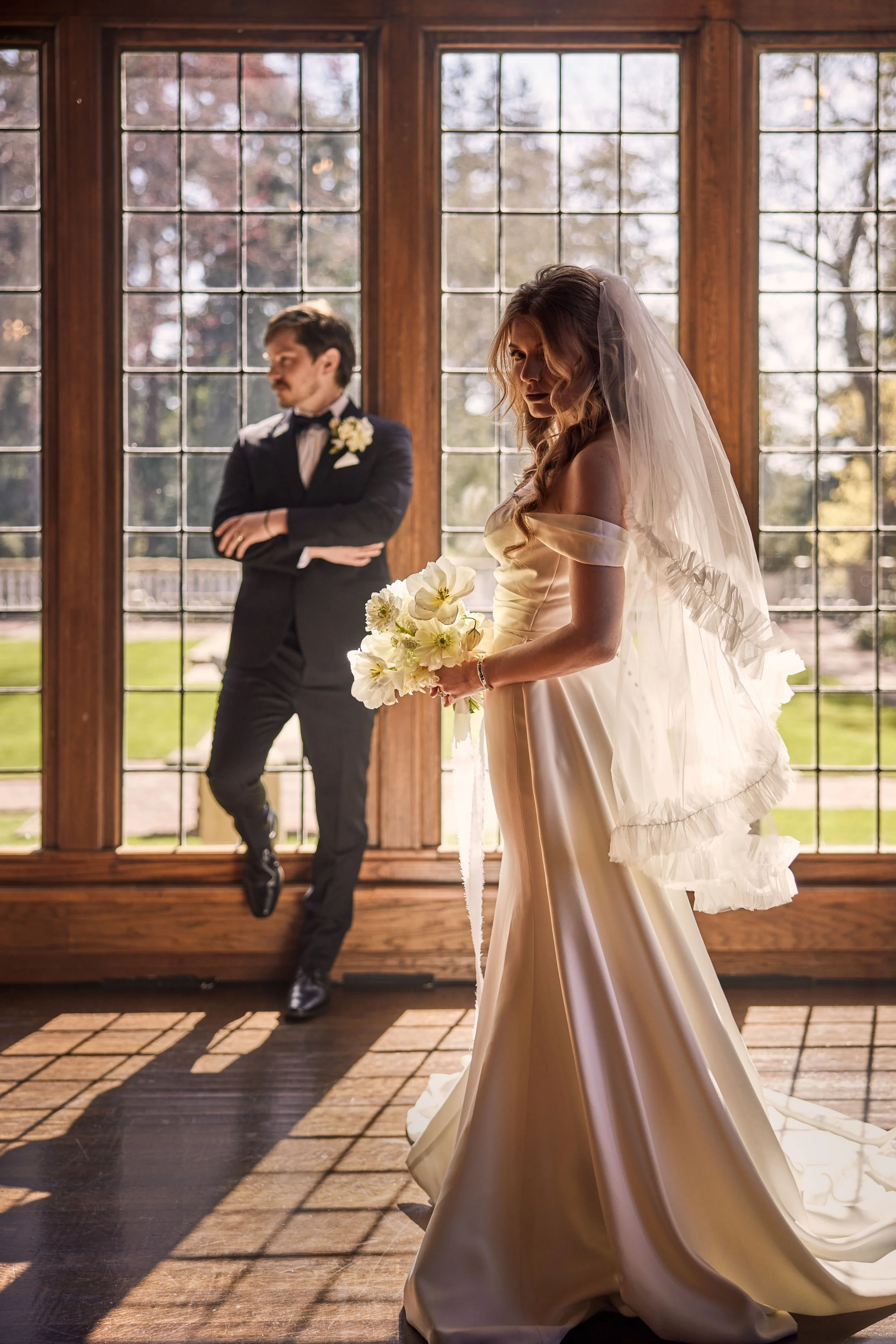 Bride in a satin wedding gown with off-the-shoulder sleeves holding a bouquet of white flowers, standing by a large window with wooden framing, with a groom in a tuxedo blurred in the background.