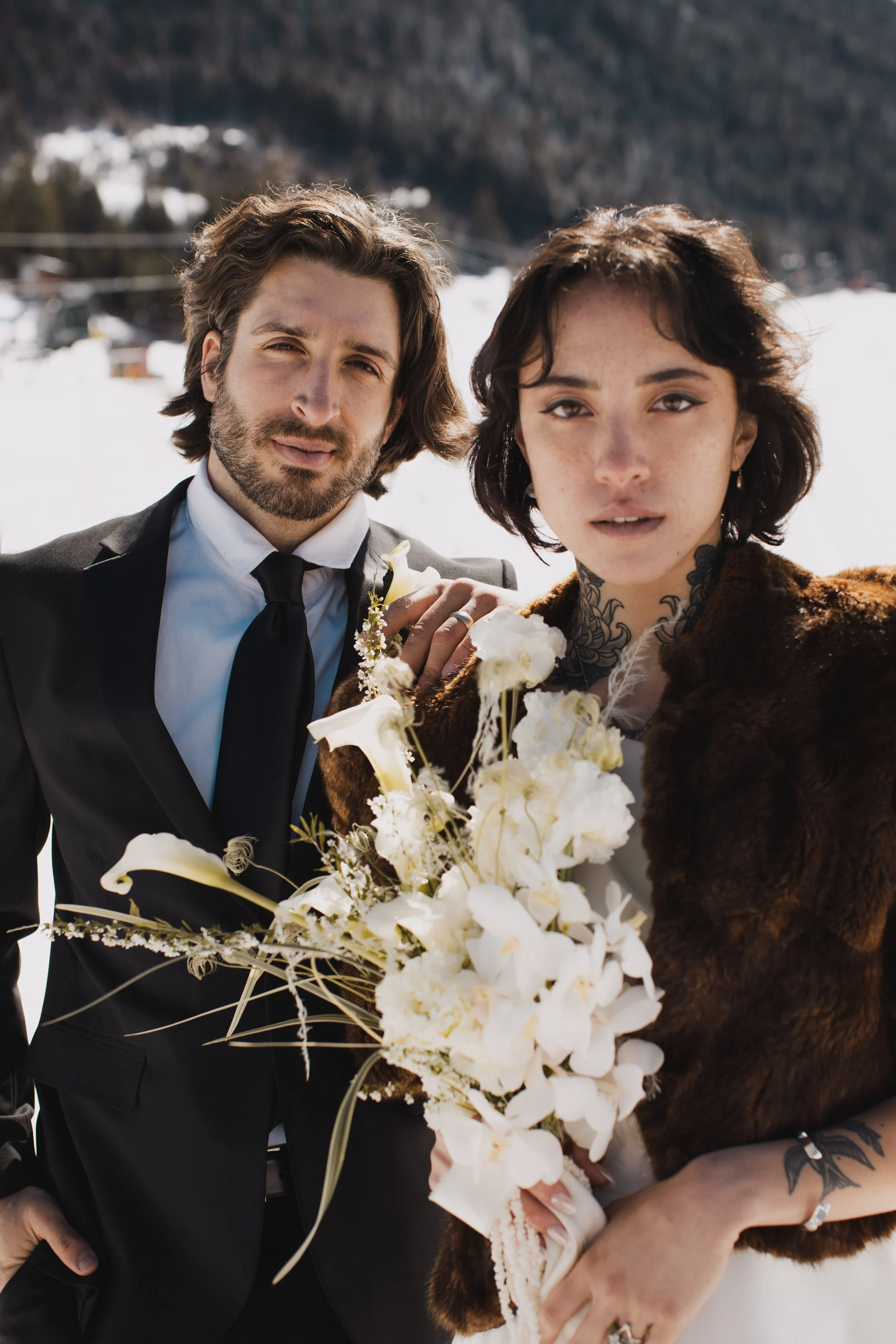 A man in a black suit and tie standing next to a woman with short dark hair, wearing a fur coat, holding a bouquet of white flowers with a snowy mountain background.