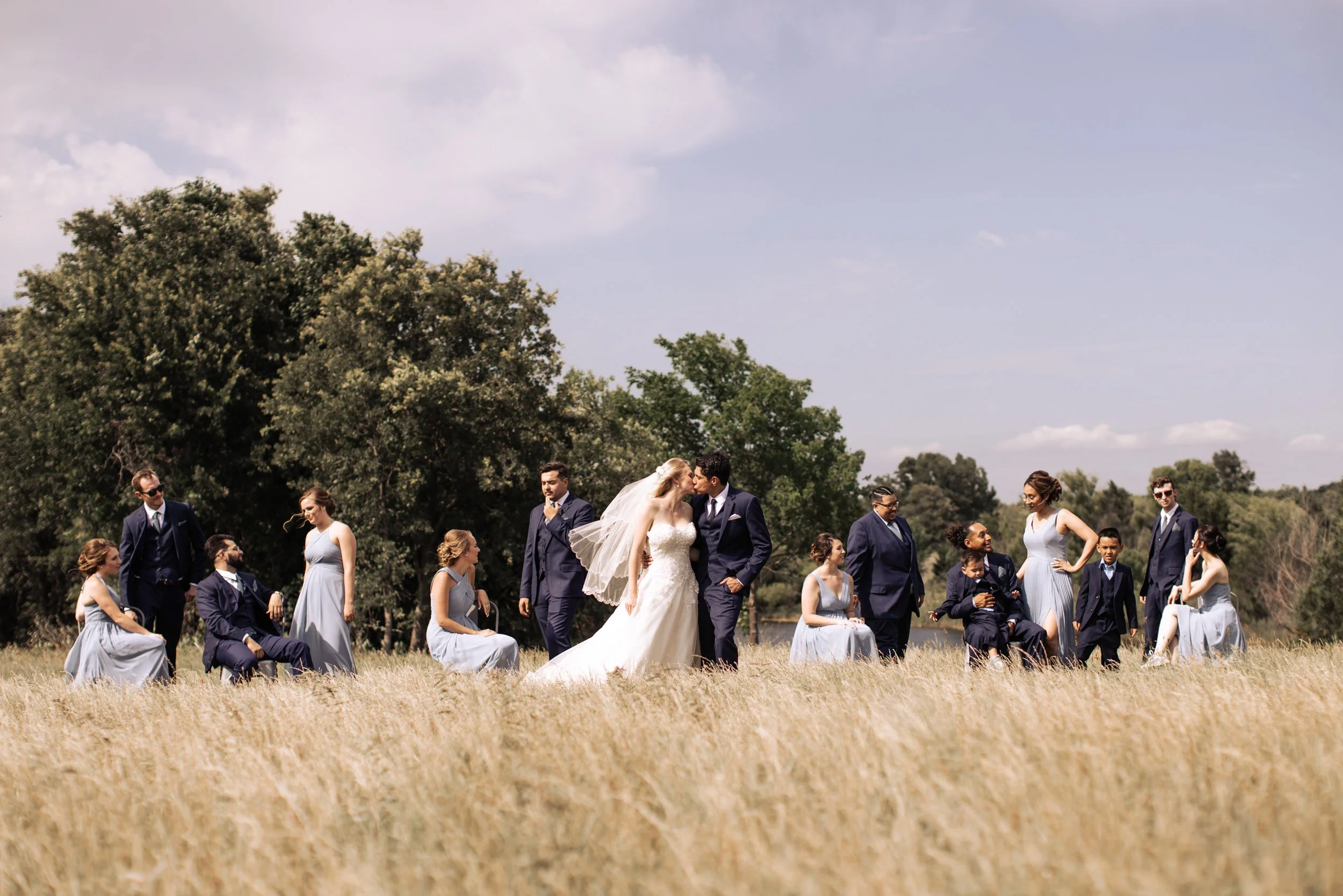 A bride and groom kiss in a field surrounded by wedding party members, with women in light blue dresses and men in navy suits, under a partly cloudy sky.