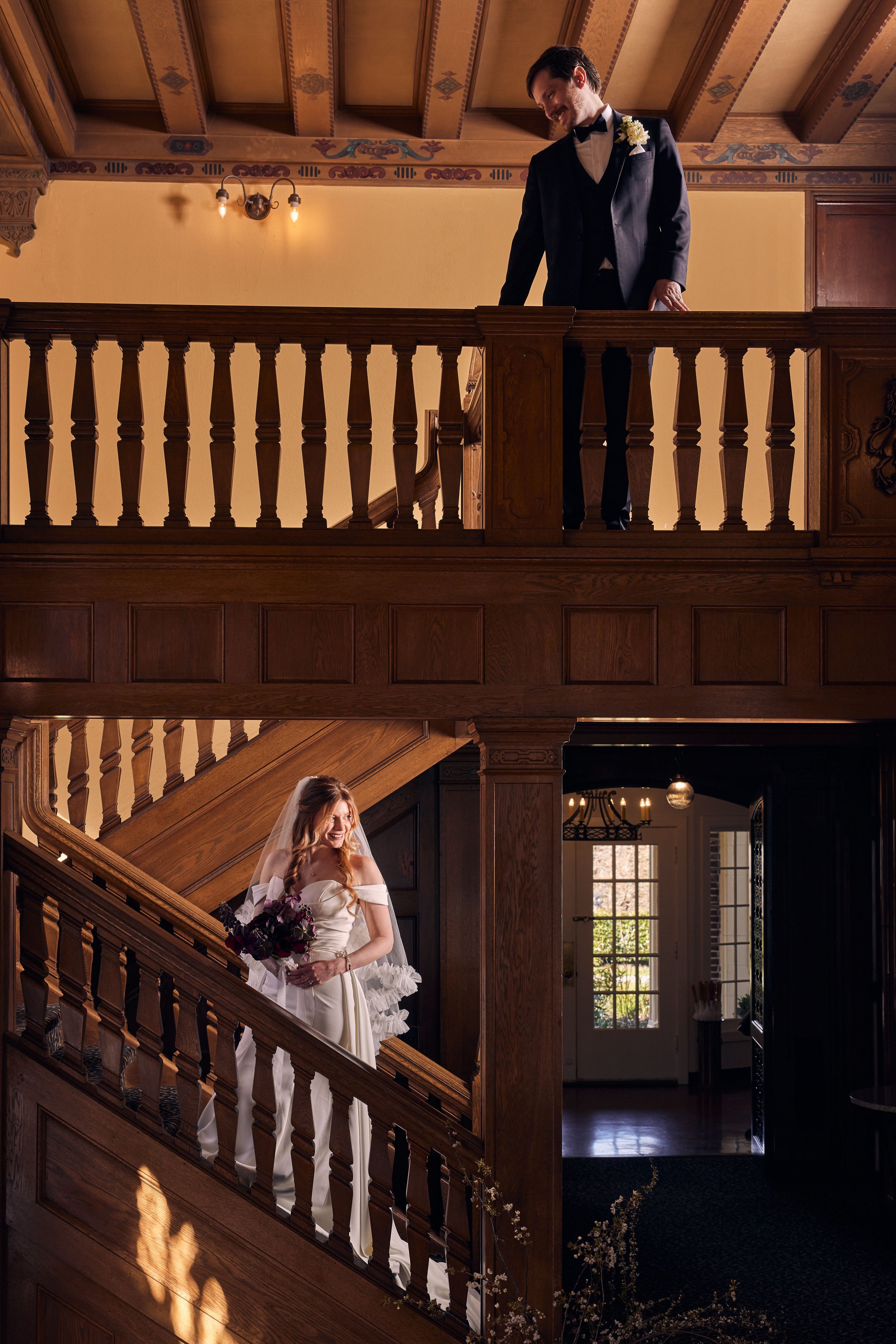 A bride in a wedding gown holding a bouquet, standing on a wooden staircase in a warmly lit, elegant interior.