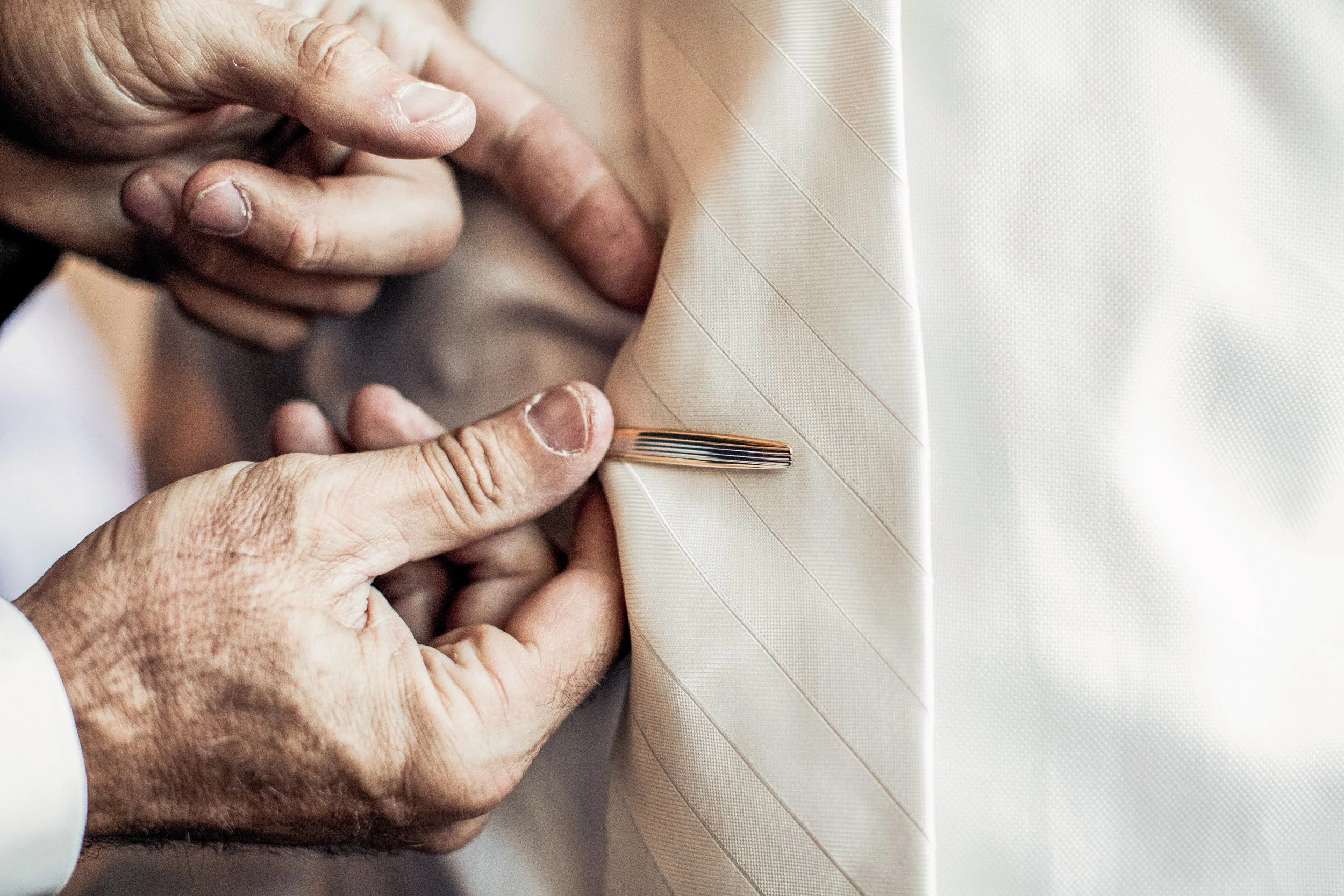 Close-up of a person adjusting a cream-colored striped tie with a tie pin.