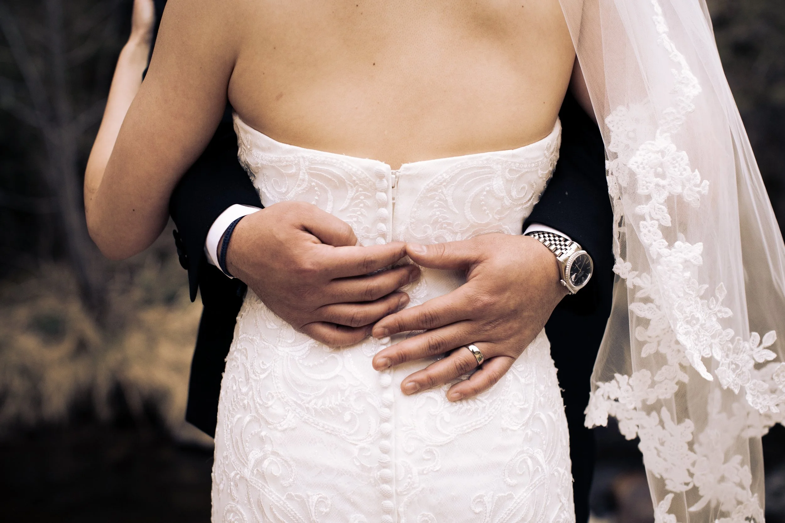 Close-up of a bride and groom holding each other, with the focus on their hands and the back of the bride's white wedding dress, with a lace veil visible on the right side.