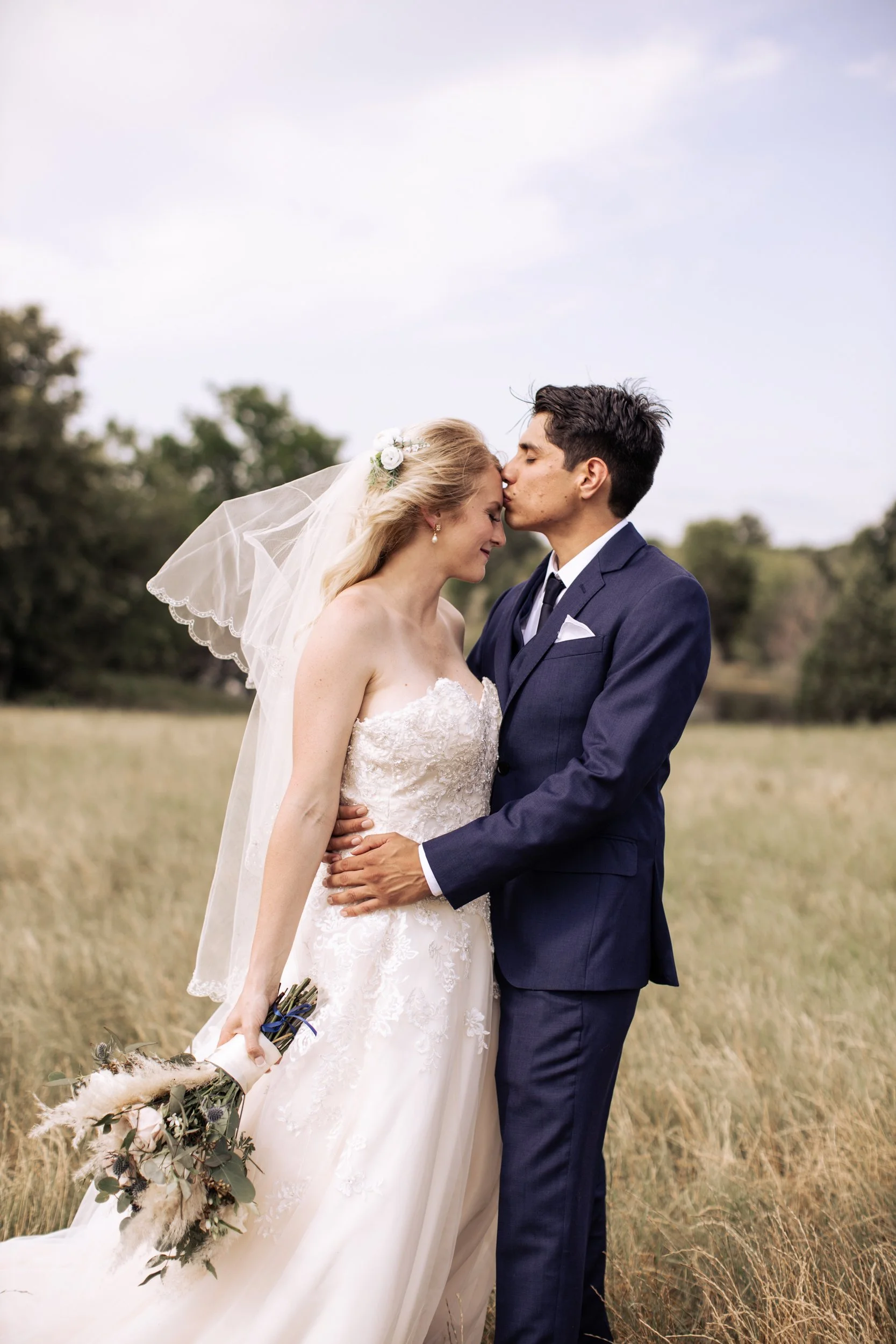 A bride and groom sharing a kiss in a field, with the bride in a strapless white wedding dress and veil holding a bouquet, and the groom in a navy suit, during their wedding photoshoot.