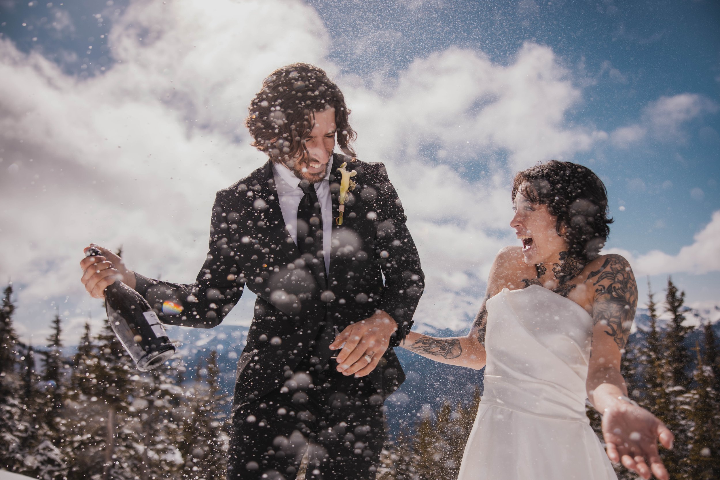 Two men, one in a tuxedo and the other in a white dress, celebrating with snow and champagne outdoors on a snowy day.