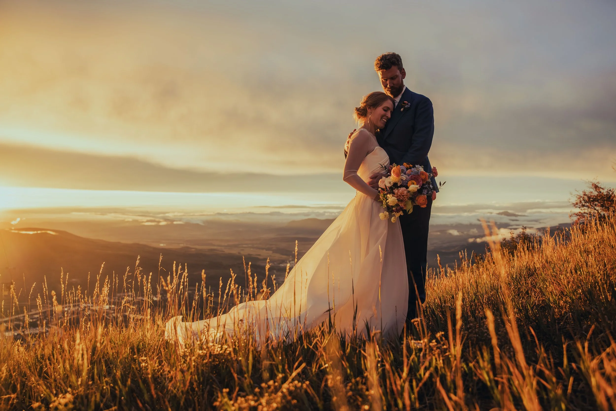 A newlywed couple embraces in a grassy field during sunset, with the bride holding a bouquet of colorful flowers and wearing a wedding dress, while the groom wears a dark suit.