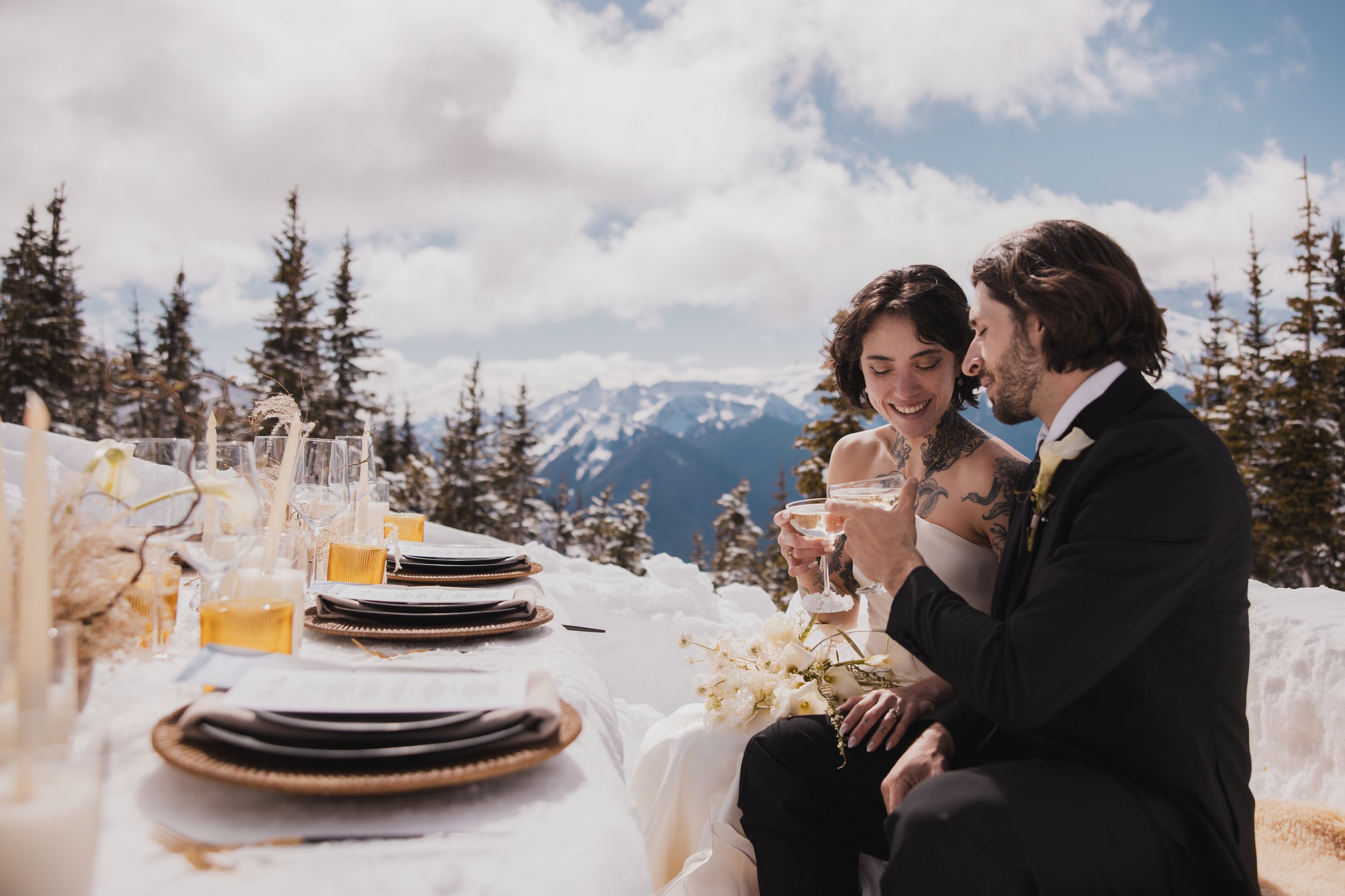 A bride and groom sitting at a long outdoor wedding table in a snowy mountainous landscape, holding glasses and smiling.