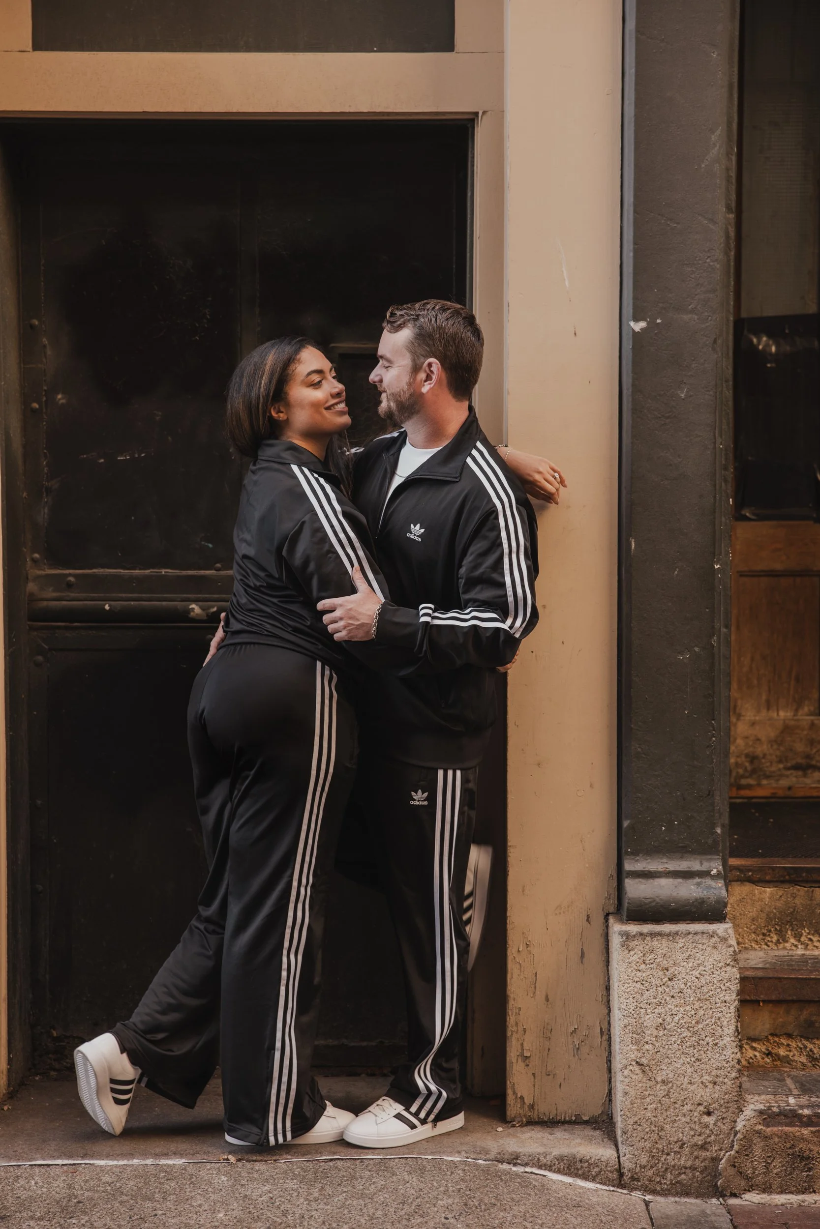 A couple in matching black Adidas tracksuits standing close and embracing each other on a city sidewalk, looking into each other's eyes.
