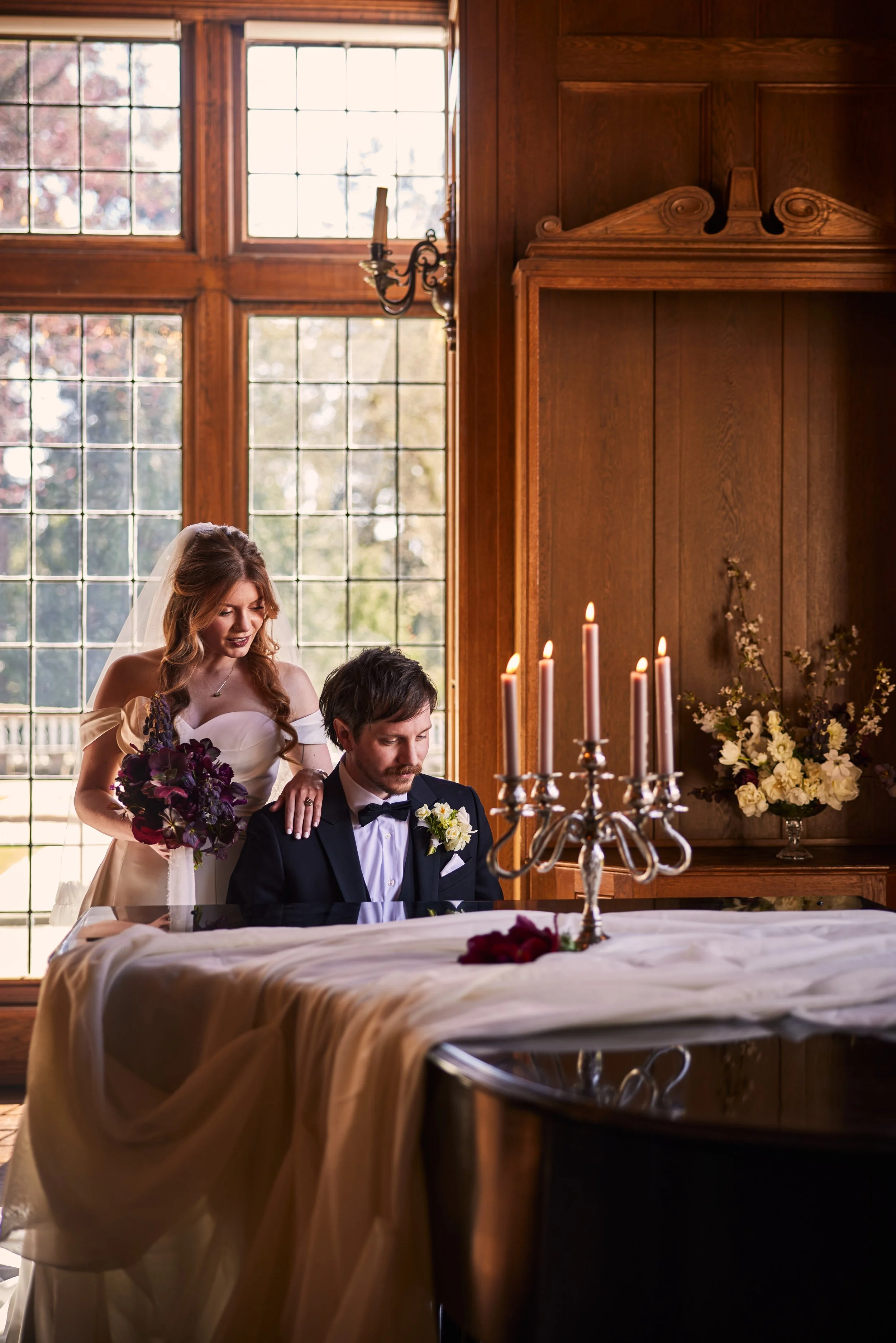 A bride and groom are participating in a wedding ceremony indoors, with large windows in the background and a wooden interior. The bride is holding a bouquet and is standing beside the seated groom, who is dressed in a tuxedo with a white shirt and b