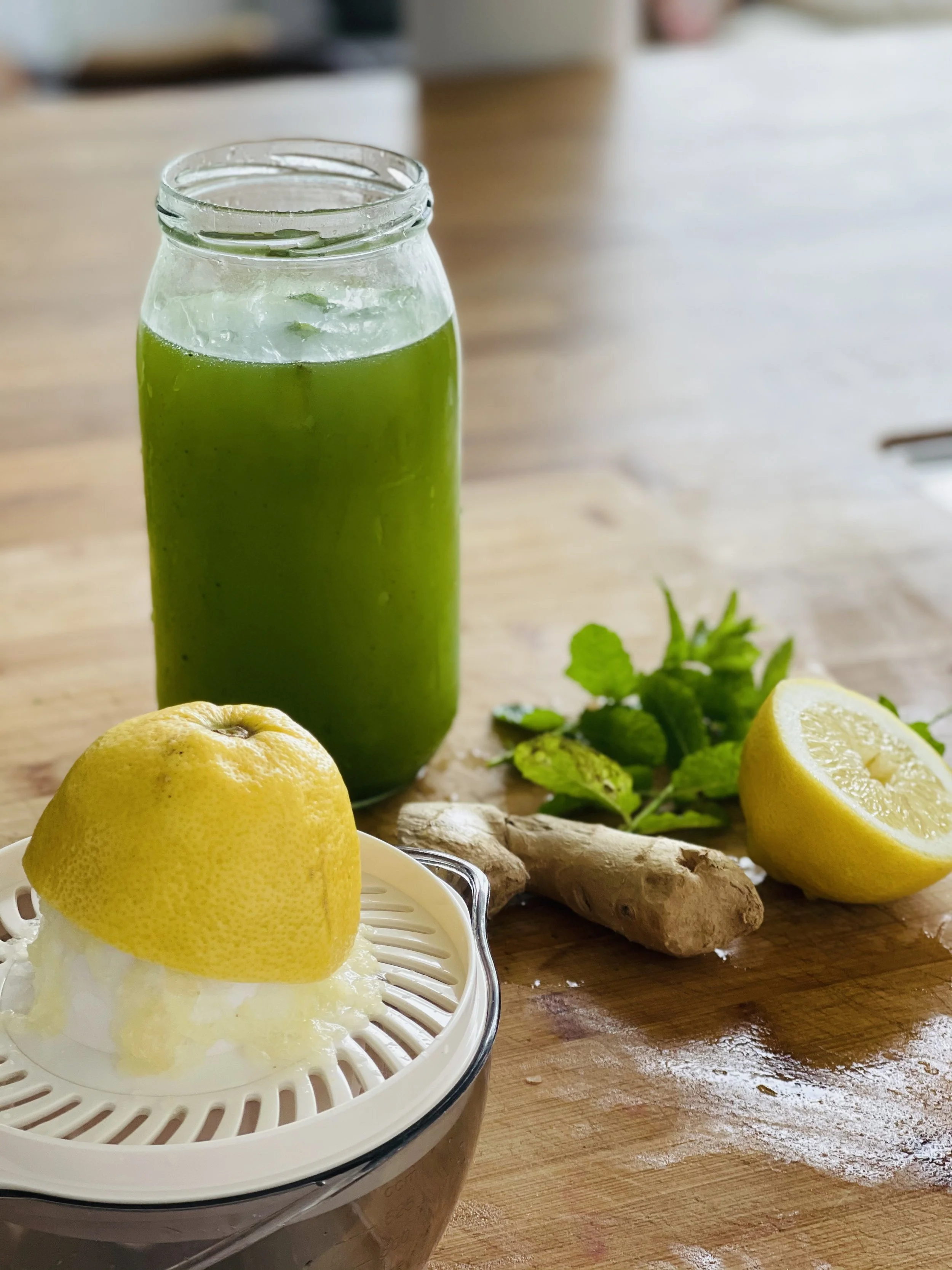 Glas met groene smoothie, citroenhelften, gemberwortel, muntblaadjes, en een citruspers met citroenhelften.