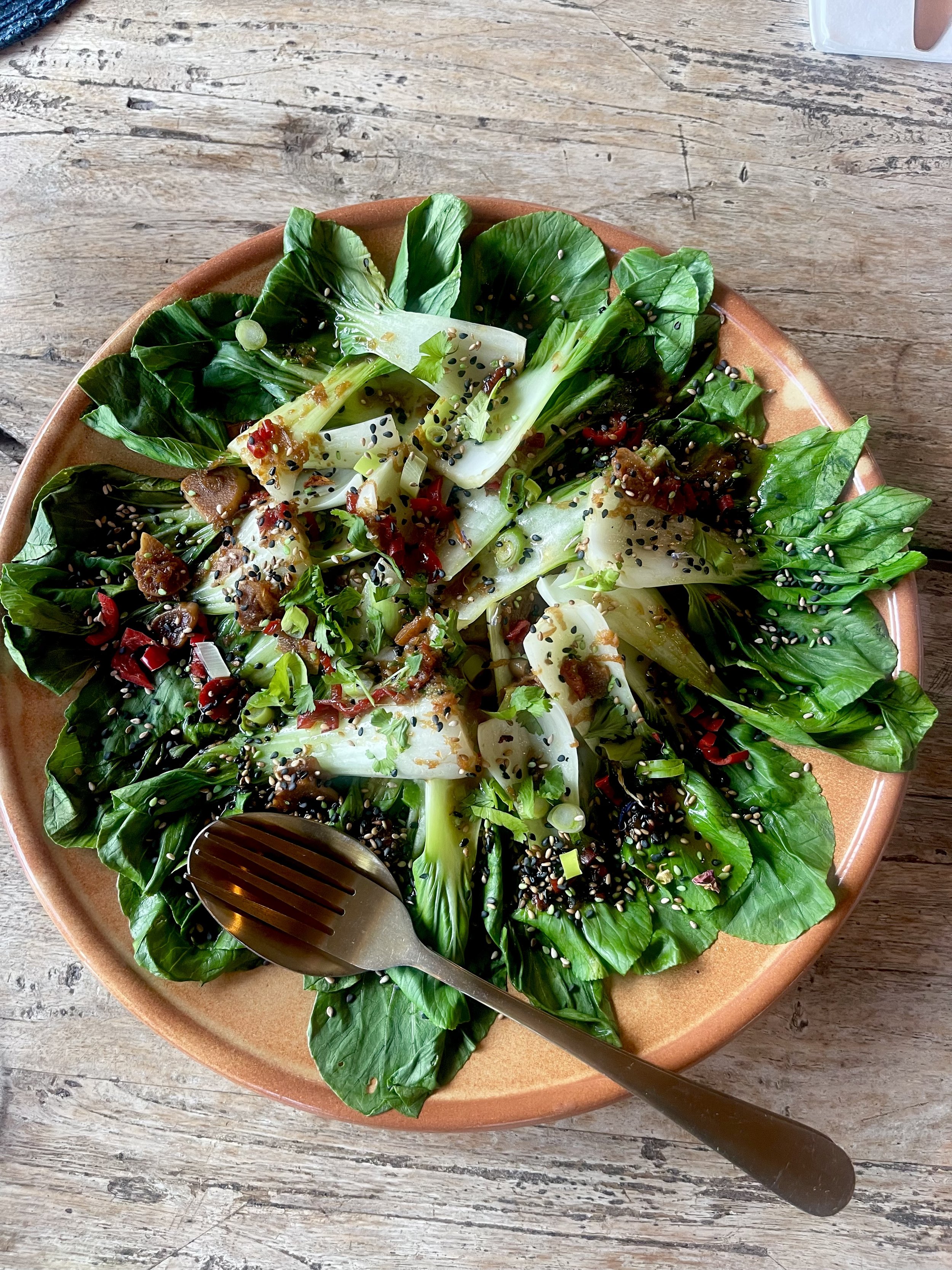 Een salade met groene bladeren, gesneden groente, sesamzaadjes, en een vinaigrette op een oranje bord, op een houten tafel.