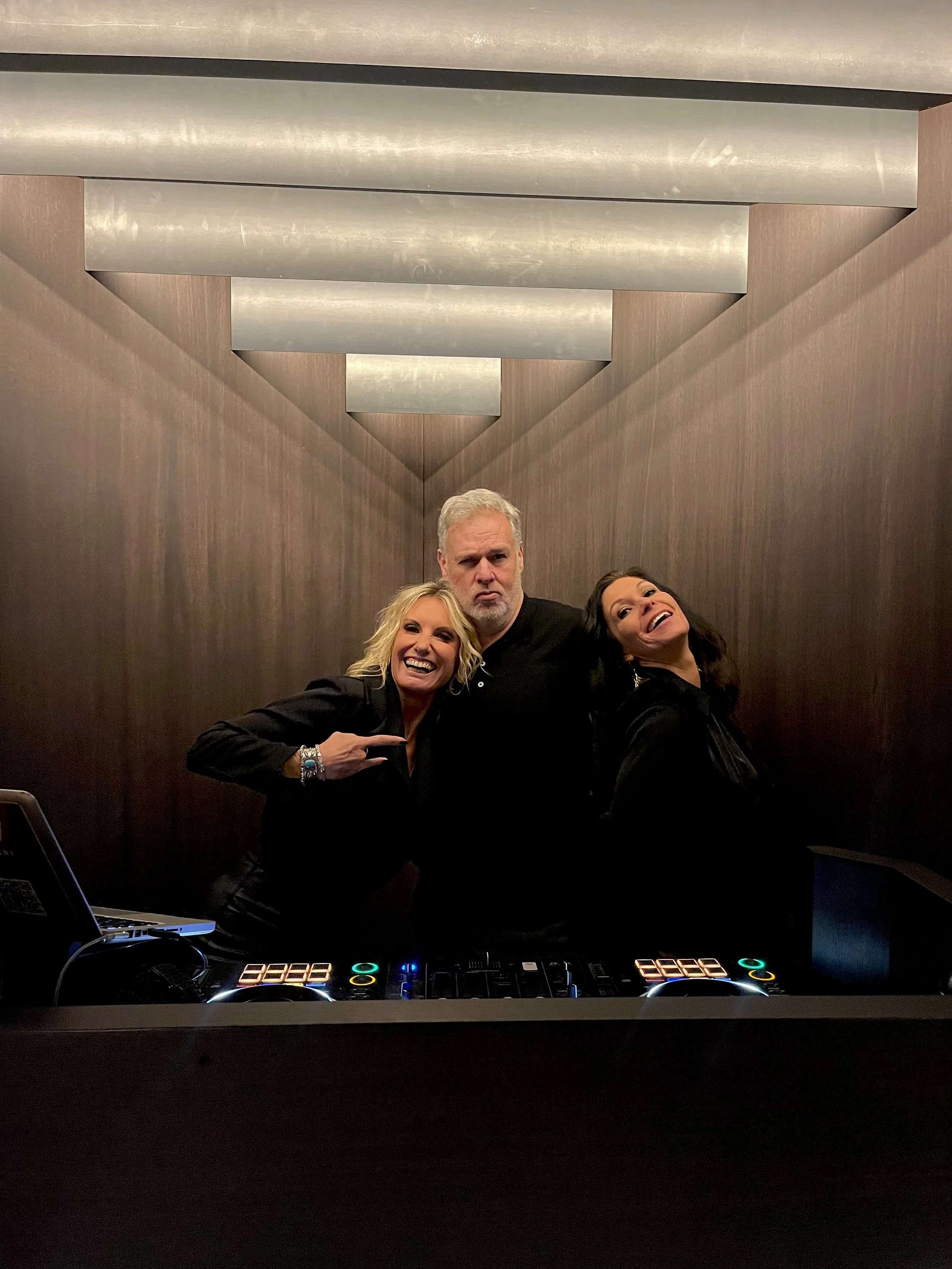 Drie mensen staan achter DJ-gear in een ruimte met een houten paneelachtergrond en metalen plafond, lachen en poseren voor de camera.