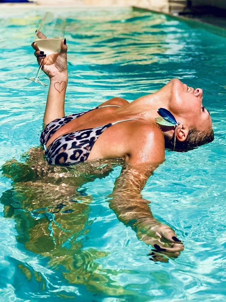 Vrouw ligt in een zwembad met een leopardprint bikini, heldere blauwe water, glas wit wijn in haar hand, grote blauwe oorbellen, opvallend donker nagellak, een hartgetekend op haar onderarm.