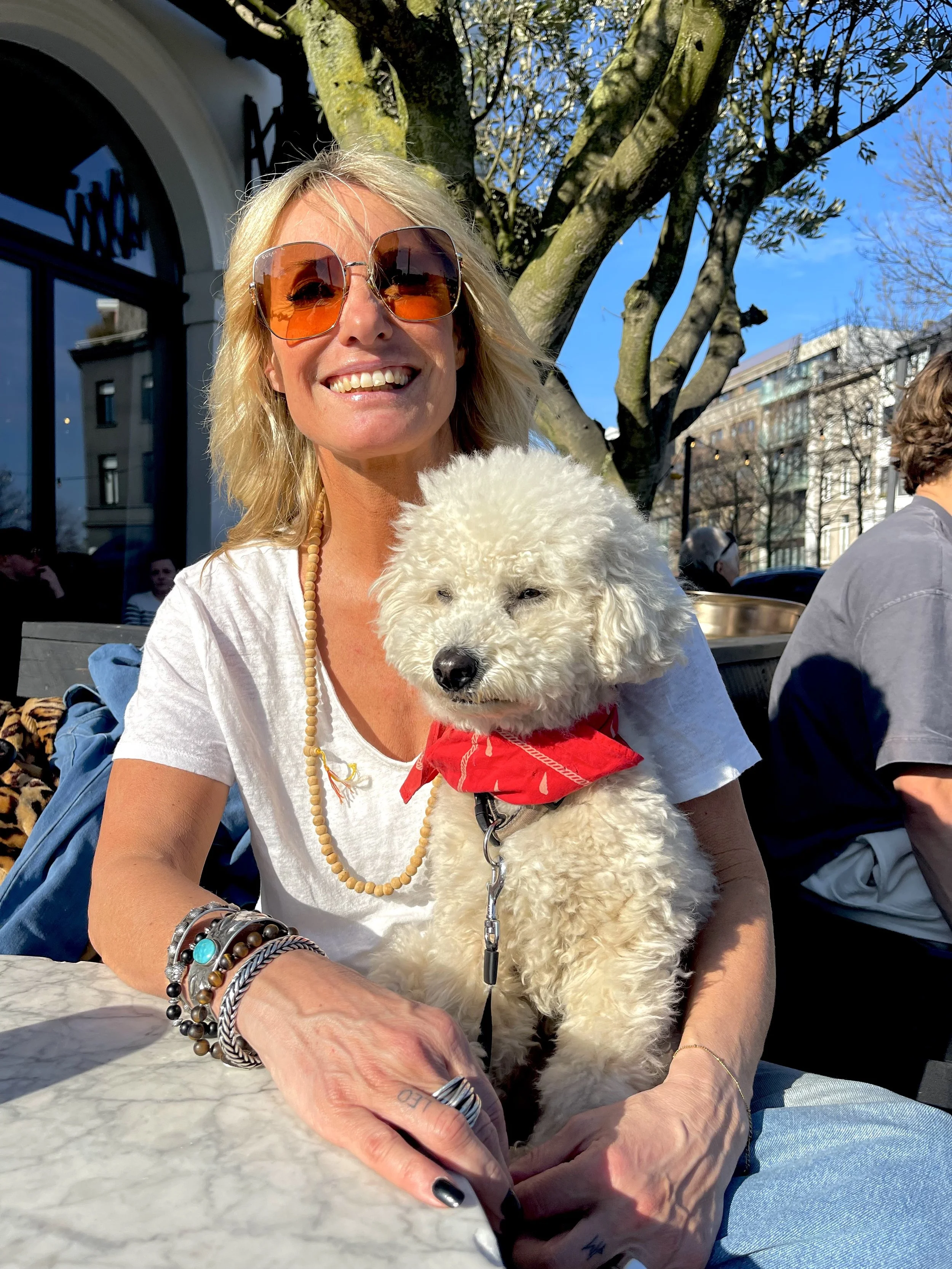 Vrouw met blonde haar, grote bruin/oranje zonnebril en meerdere sieraden, zit buiten met haar witte, donzige hond met rode bandana, onder een boom met een blauwe lucht op de achtergrond.