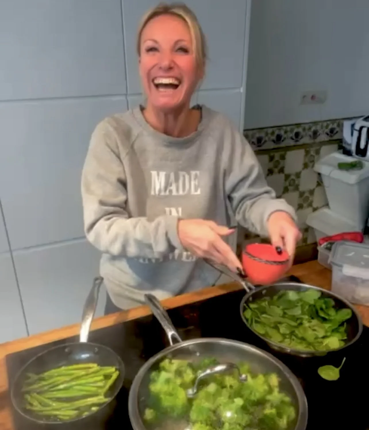 Vrouw die in een keuken groenten kookt en lachend een rood bakje vasthoudt.