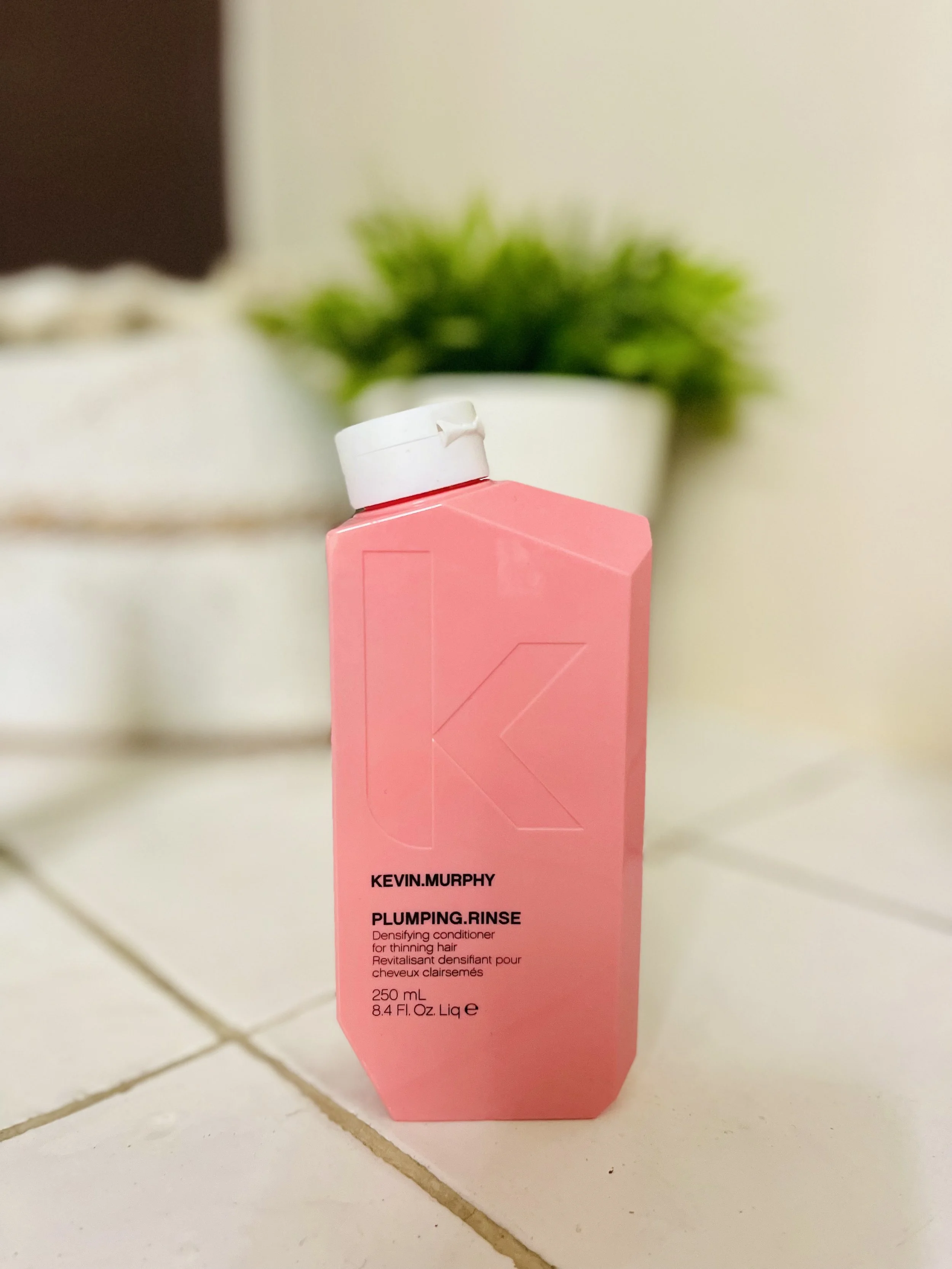 Een flesje van Kevin Murphy Plumping Rinse, een verdikkingsconditioner voor dun haar, op een witte tegeltuin, met een groen plantje op de achtergrond.
