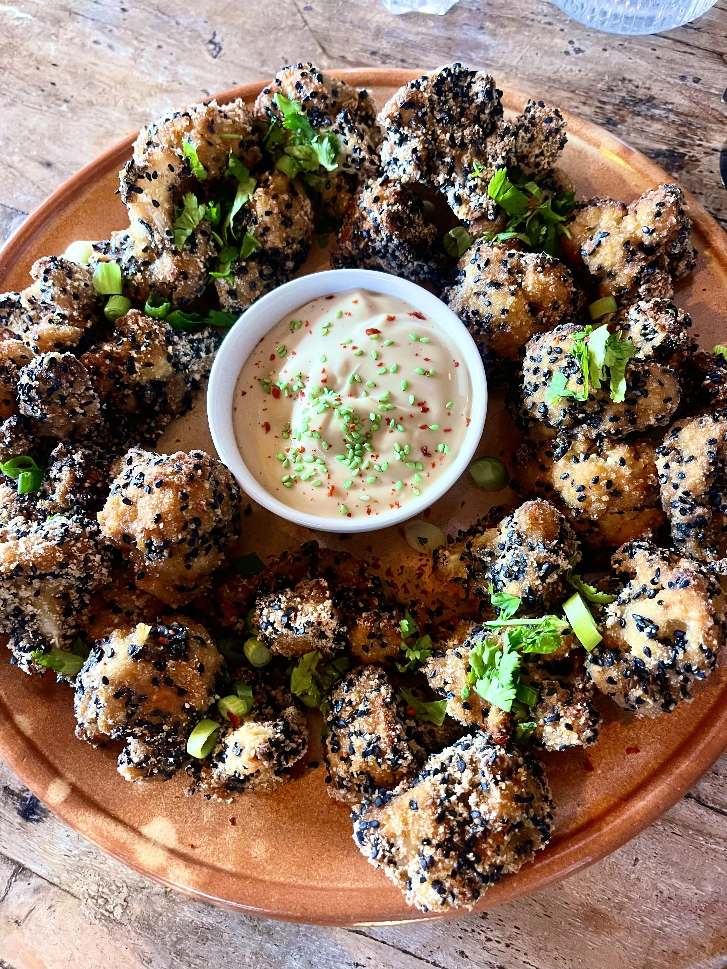 Gefrituurde stukjes kip met sesamzaad en koriander, geserveerd met een saus in een klein wit kommetje.