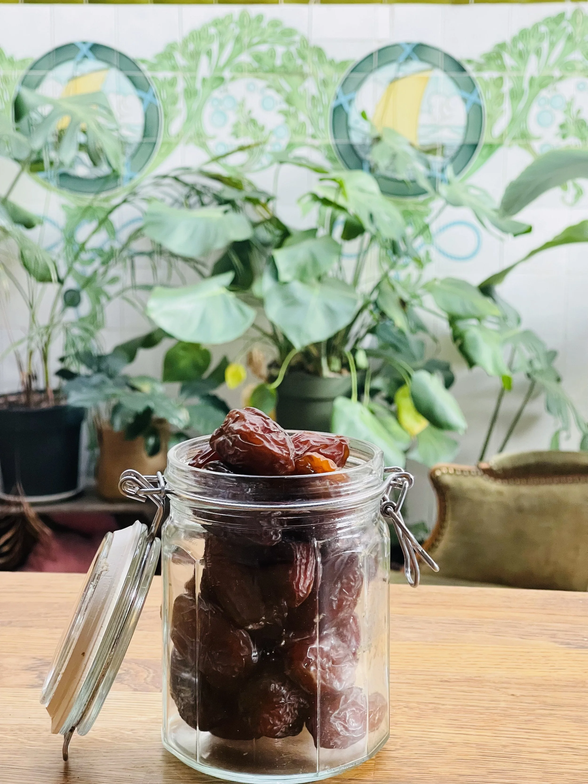 Een glazen pot met datum dadels op een houten tafel, met groene planten op de achtergrond.