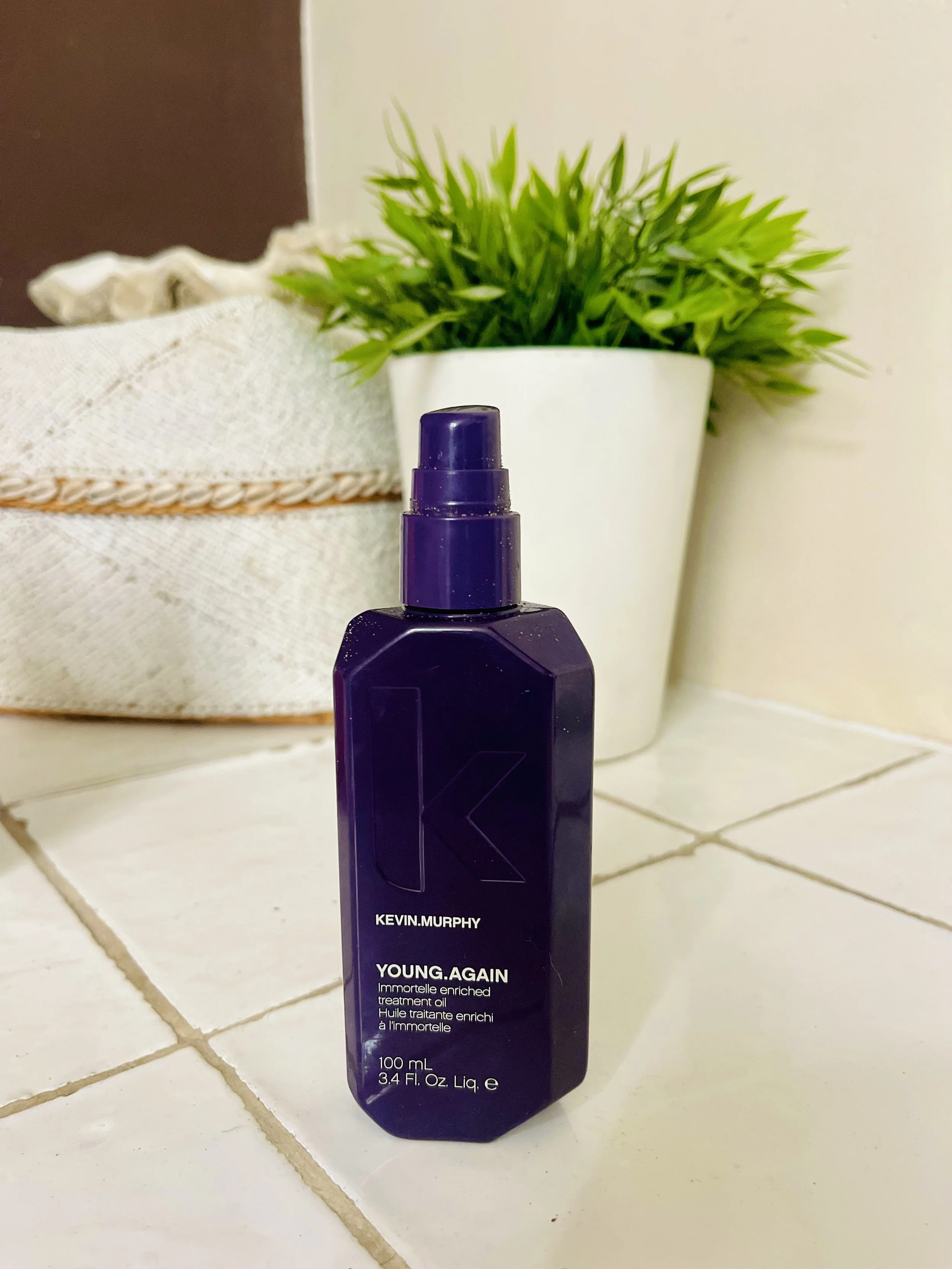 Een fles Kevin Murphy Young Again behandeling olie op een wit betegeld oppervlak naast een bloempot met groen plant en een mand met doek.