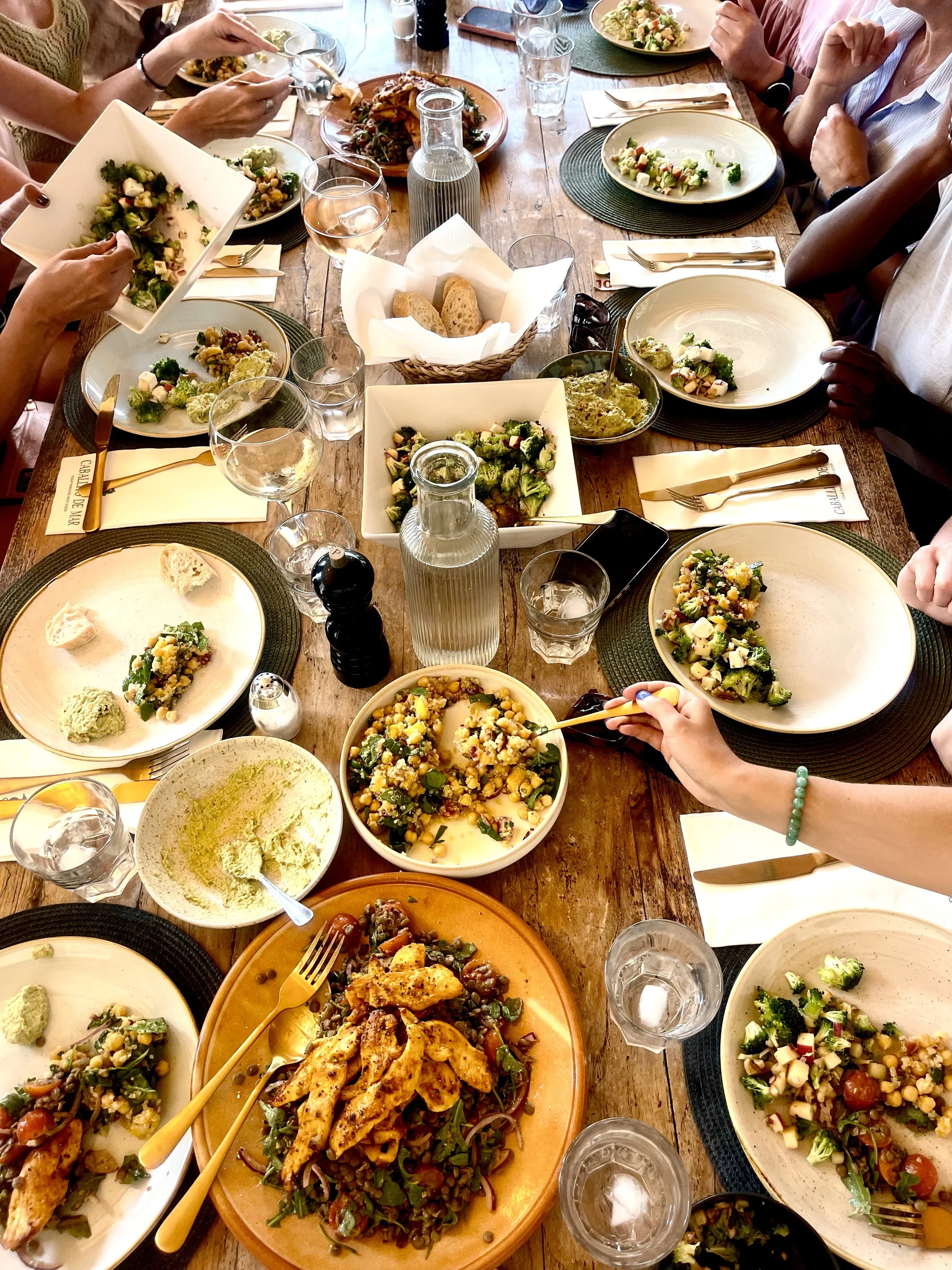 Een grote eettafel met diverse salades, een bord met gegrilde kip, brood en glazen water en wijn, omgeven door mensen die samen eten.