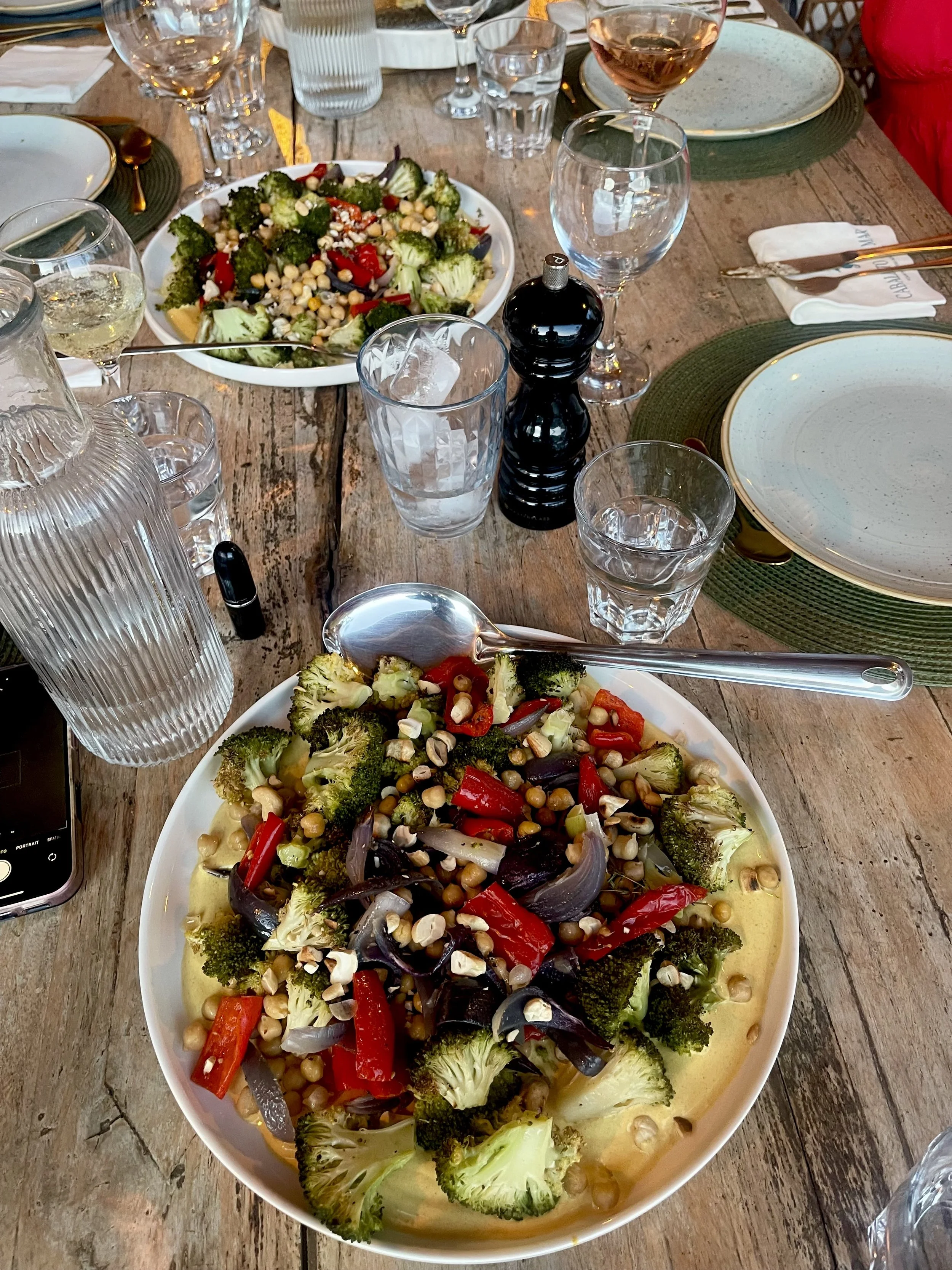 Een grote schaal met een salade van broccoli, rode paprika, gekookte kikkererwten en linzen, geserveerd op een houten tafel met glazen en bestek.