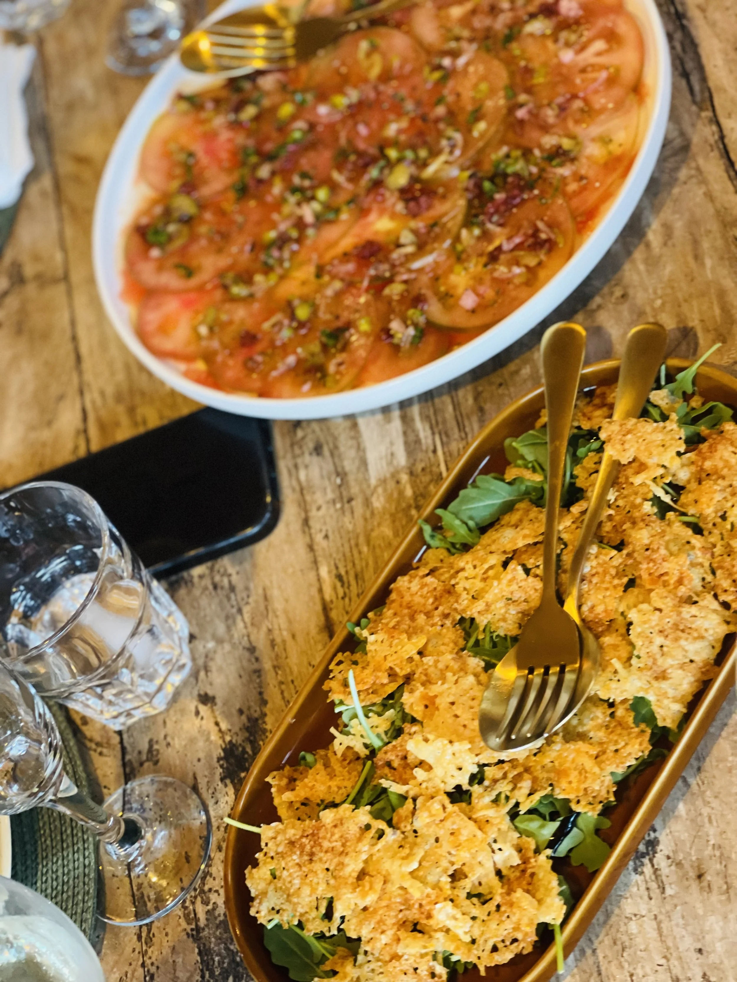 Gerecht met tomatensalade met gesneden tomaten, ui en kruiden naast een schaal met gefrituurde kip op een bedje van greens, op een houten tafel.
