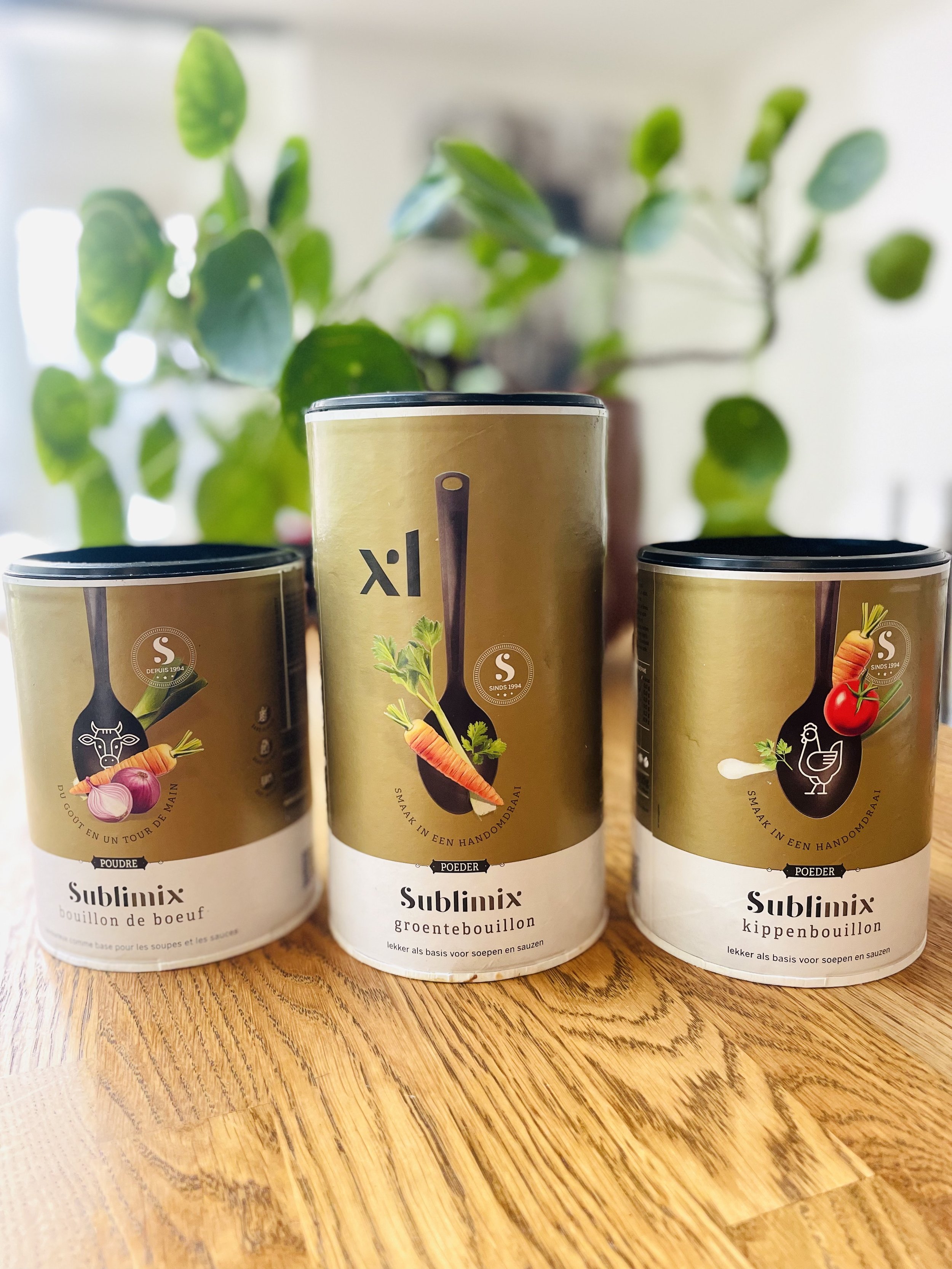 Drie blikjes Sublimix groentebouillon en kippenbouillon op een houten tafel, met een groene plant op de achtergrond.