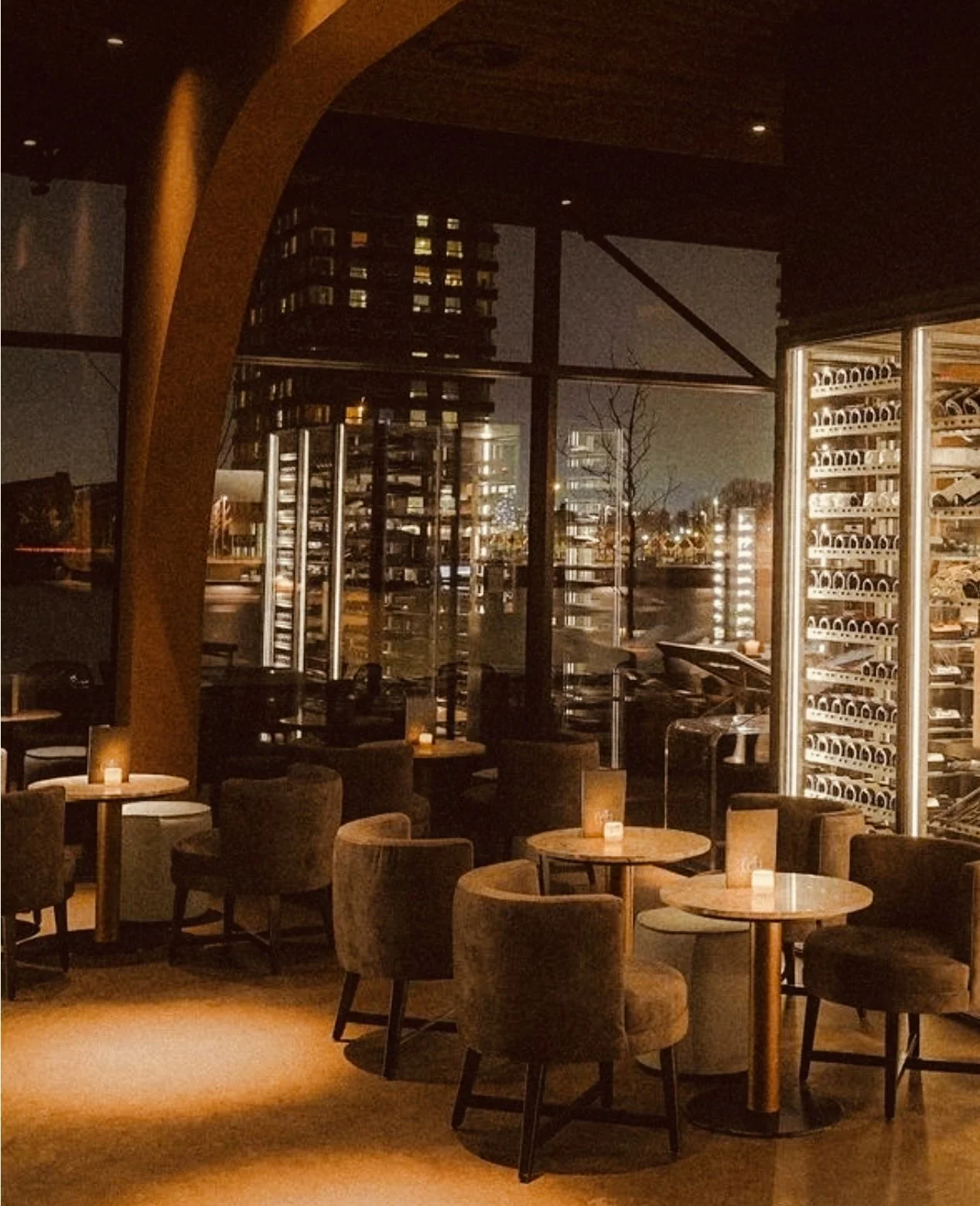 Gezellige restaurant of bar met ronde tafels, thermostaten en wijnrekken, binnen met uitzicht op een stad in de avond.