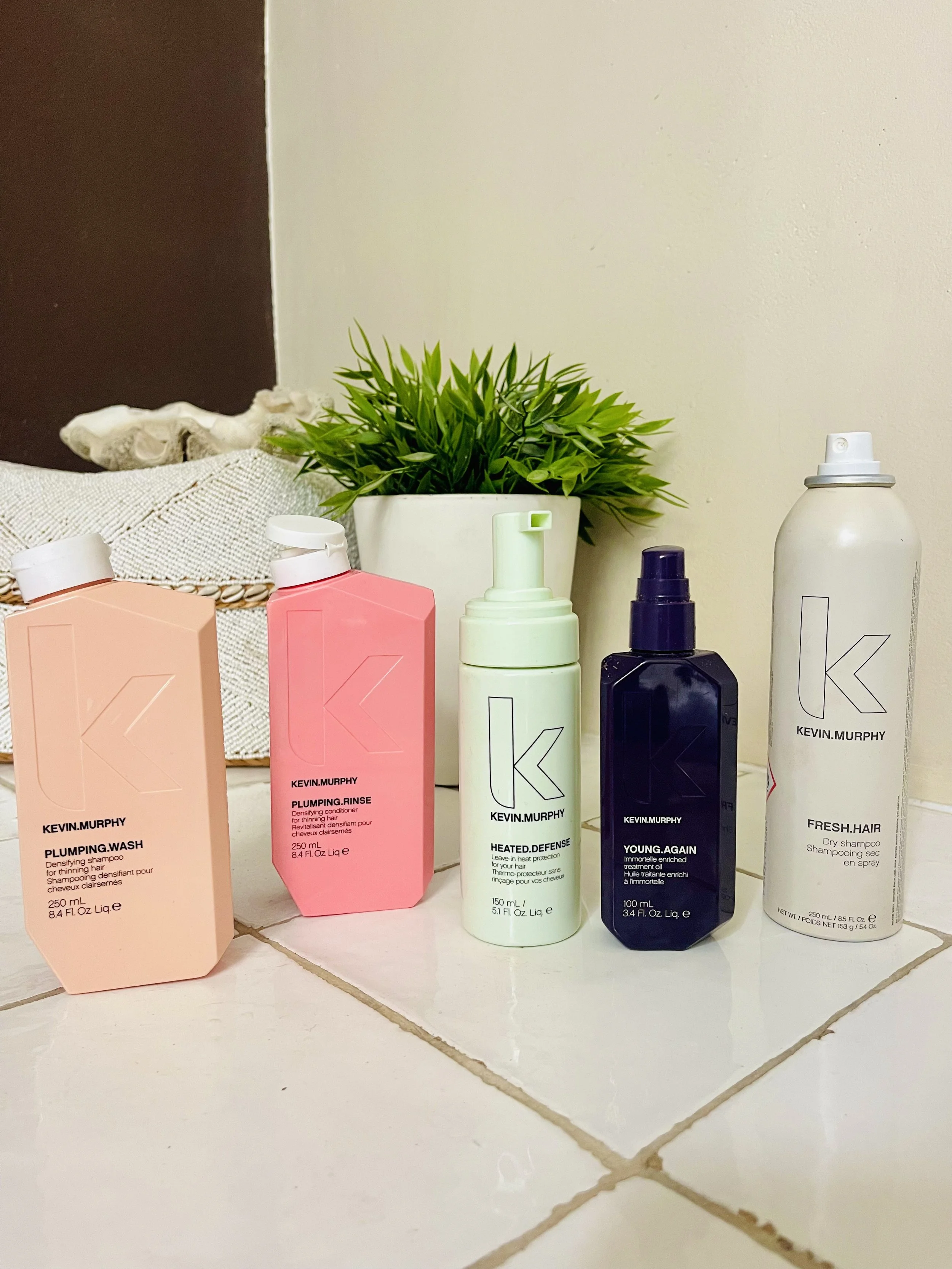 Verschillende haarverzorgingsproducten van Kevin.Murphy op een witte tegelvloer met een groen plantenpot op de achtergrond.