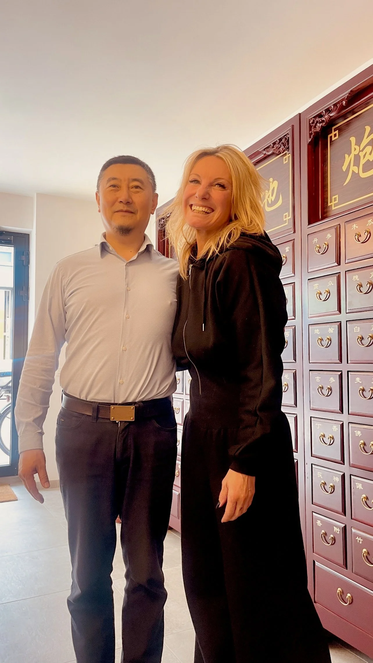 Twee mensen staan naast elkaar binnen, voor een muur met Chinese tekens en kleine laden met koperen handvatten, lachend en poserend voor de camera.