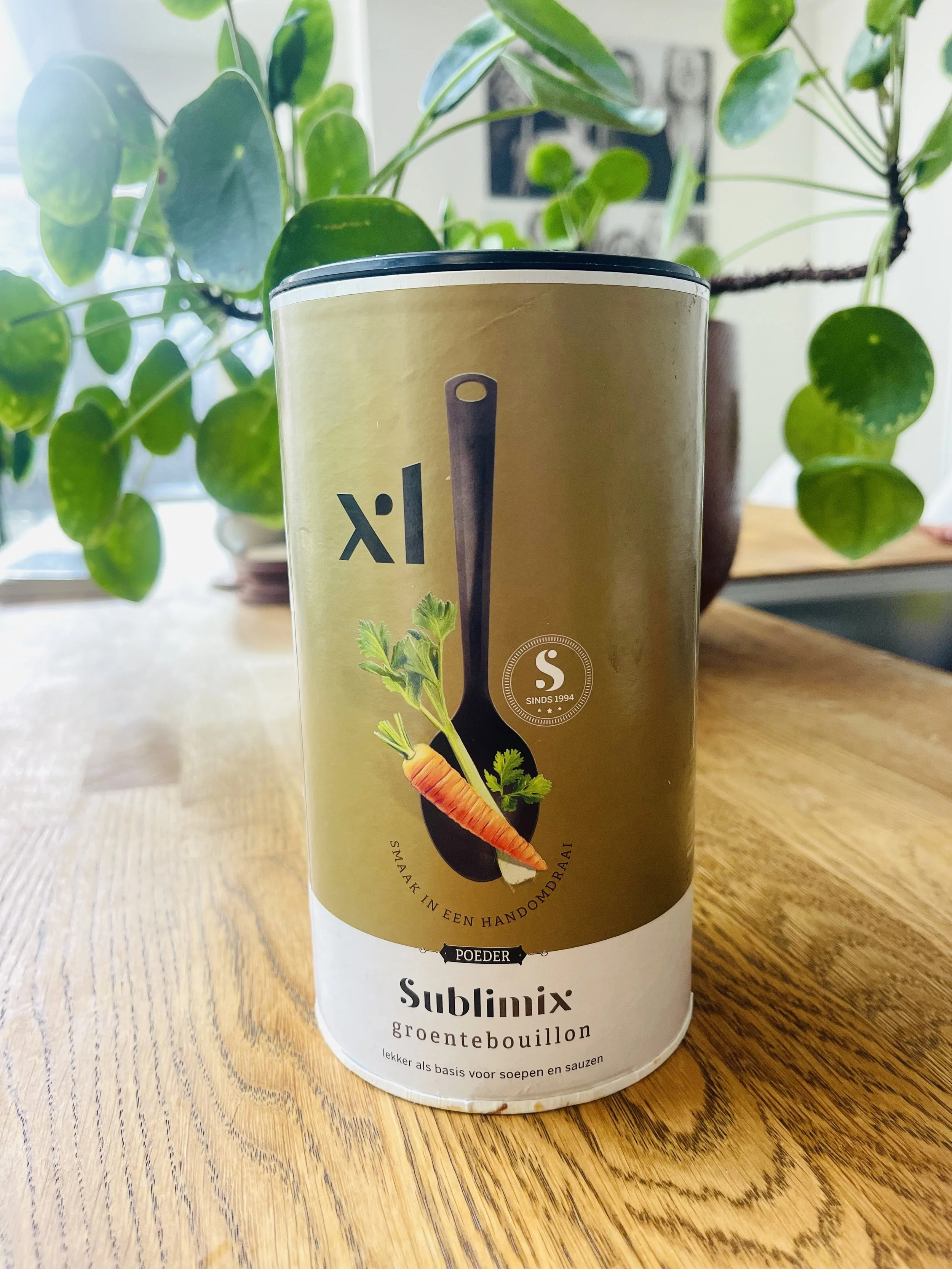 Blik op een doosje Sublimax groentebouillon met een achtergrond van groene planten op een houten tafel.