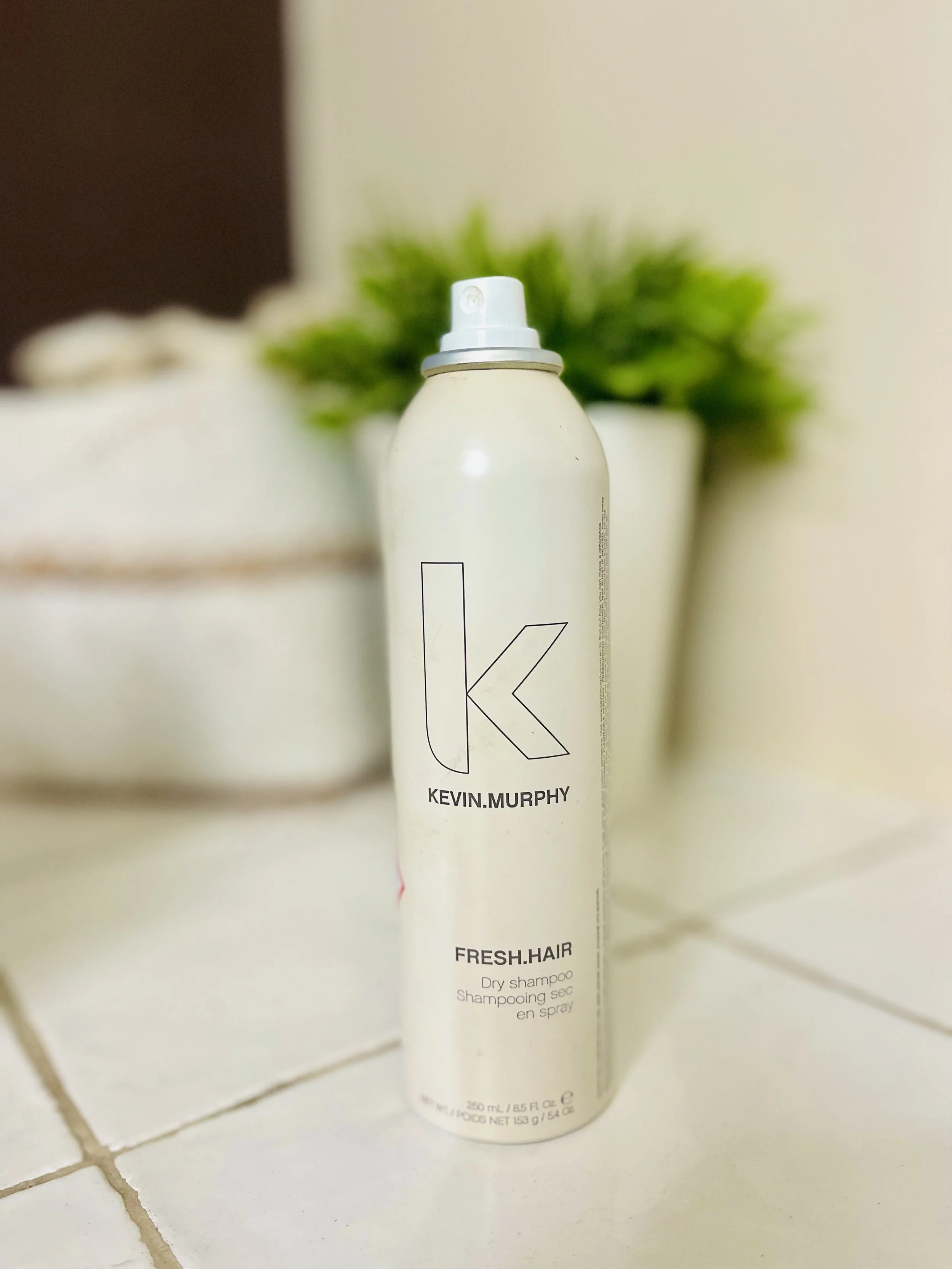 Een fles Kevin Murphy Fresh Hair droogshampoo op een badkamertafel, met een plantenbak en badhanddoeken op de achtergrond.