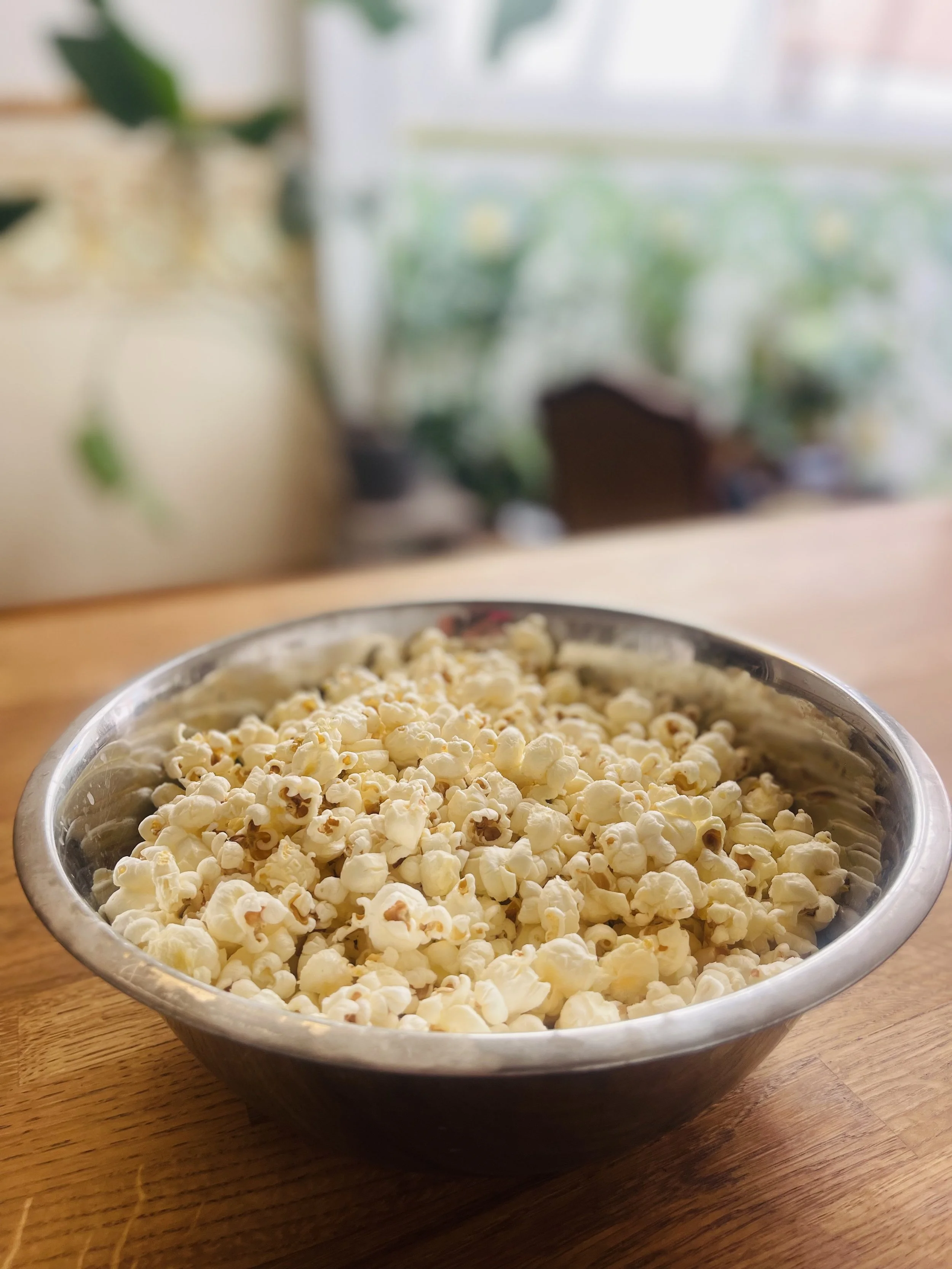 Een metallic kom gevuld met popcorn op een houten tafel, met een blurred achtergrond van een kamer met planten en een raam.