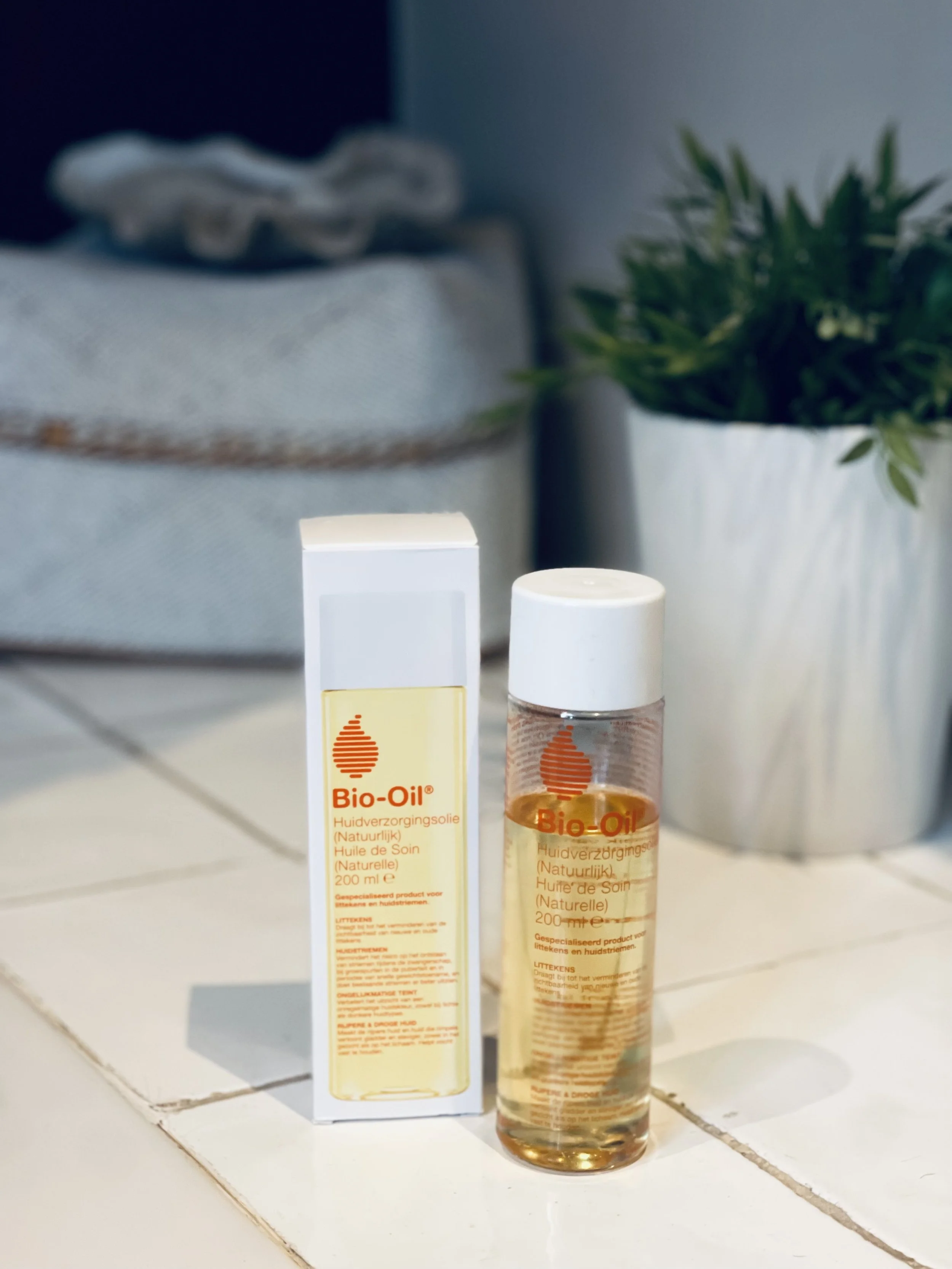 Een fles Bio-Oil huidverzorgingsolie naast de bijbehorende verpakking op een witte tafel, met een grijze en groene plant op de achtergrond.