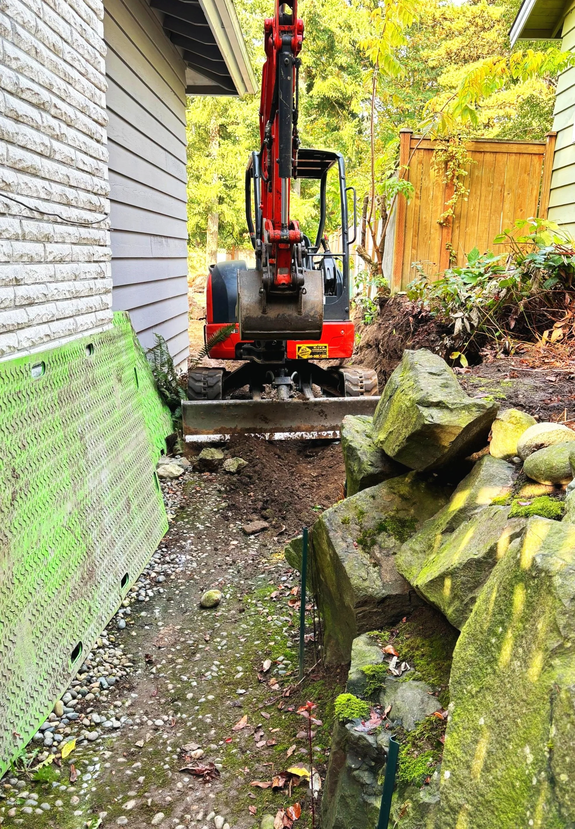 An excavator between two houses clearing boulders to build a landscaping retaining wall in Snohomish County Washington.