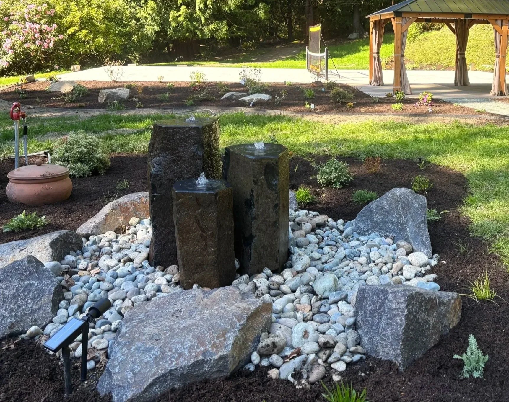 A landscaped garden with a small water fountain made of three dark stones with water spouting from the top. Surrounding rocks, pebbles, green plants, freshly mulched flower beds, a gazebo with curtains, and a tennis court can be seen in the background.