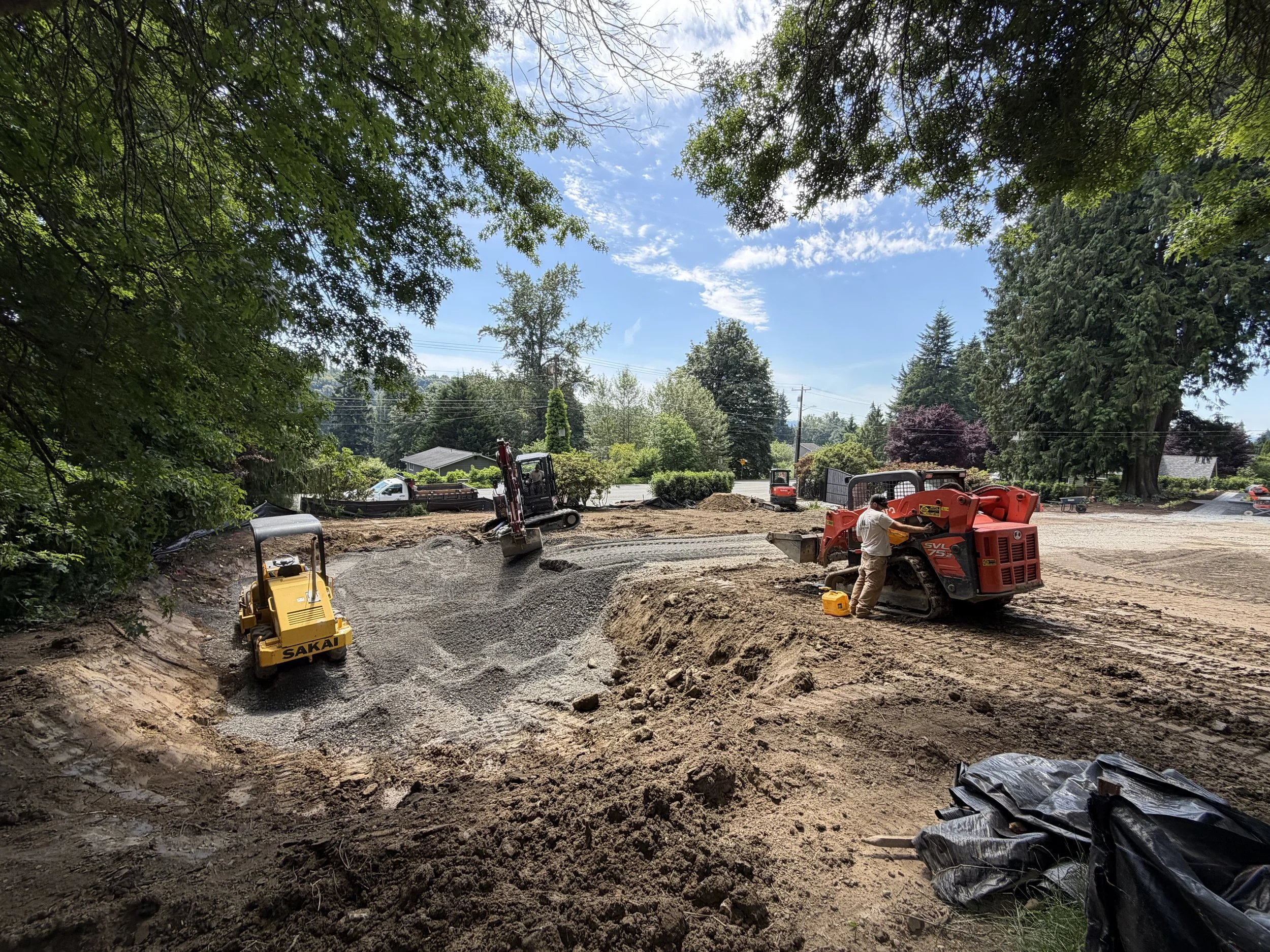 Construction site with machinery, workers, and gravel, surrounded by trees under a partly cloudy sky.