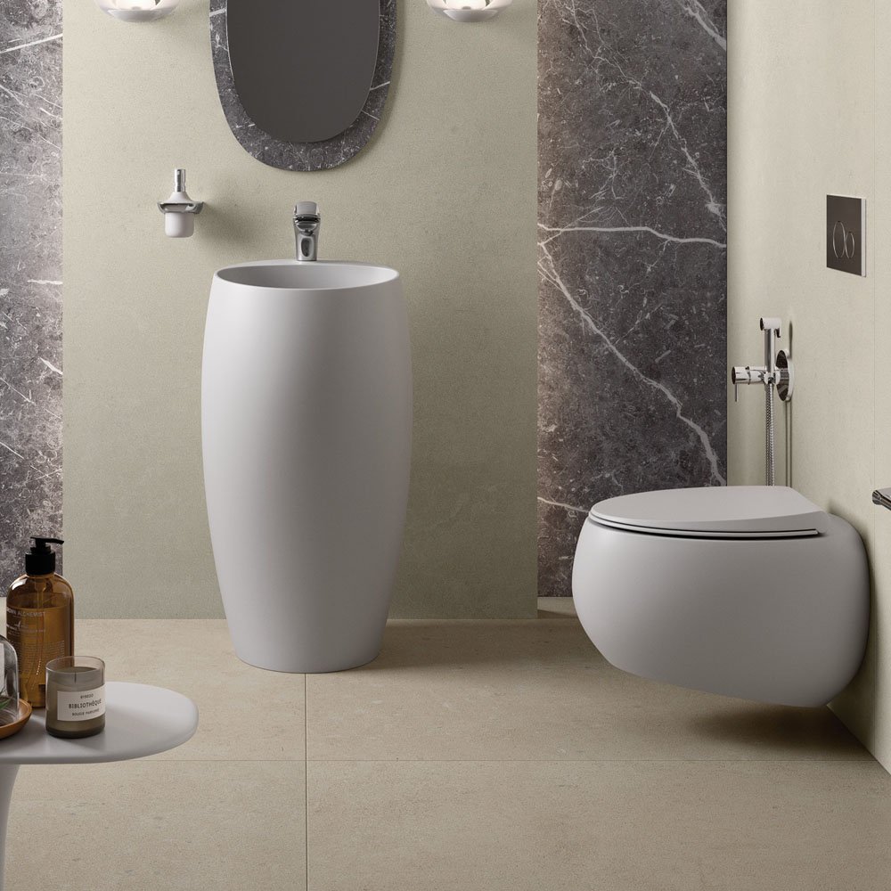 Modern bathroom interior with an oval pedestal sink, wall-mounted toilet, and a marble-accented wall. A round mirror and a small table with toiletries are visible.