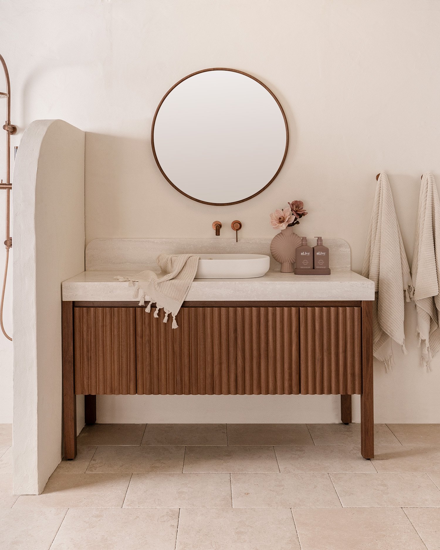 Minimalist bathroom with round mirror, wooden vanity, ceramic basin, and beige towels. Decor includes flowers and soap dispensers.