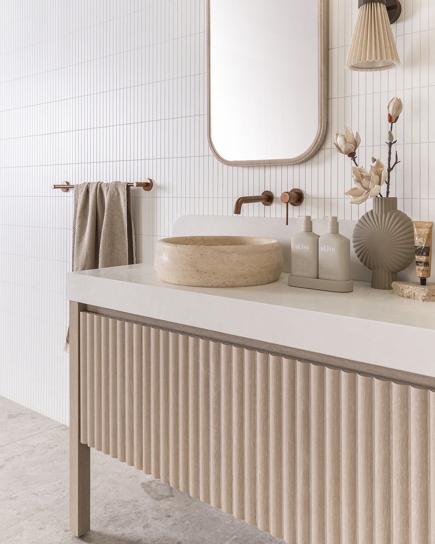 Modern bathroom with vertical white tiles, a beige stone sink, wall-mounted faucet, a rectangular mirror with rounded corners, a wall lamp with a pleated shade, beige accessories, and a wooden vanity with fluted design.