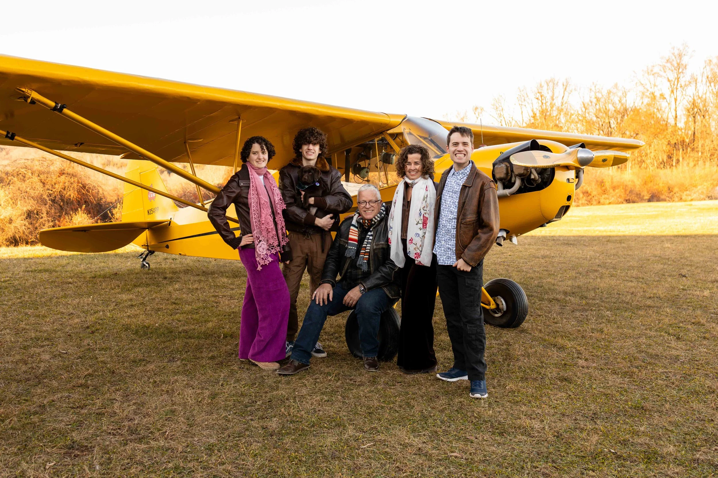 A family of five wears leather jackets and scarves while standing in front of an antique yellow plane - family portrait taken by Jennifer Fulton Photography in Central New Jersey