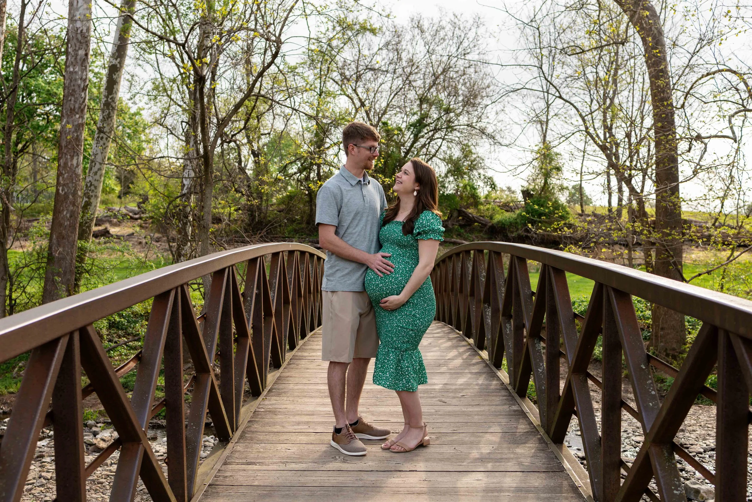 A pregnant couple stands on a bridge wearing gray, tan, and green outfits - maternity photo taken by Jennifer Fulton Photography in Central Jersey