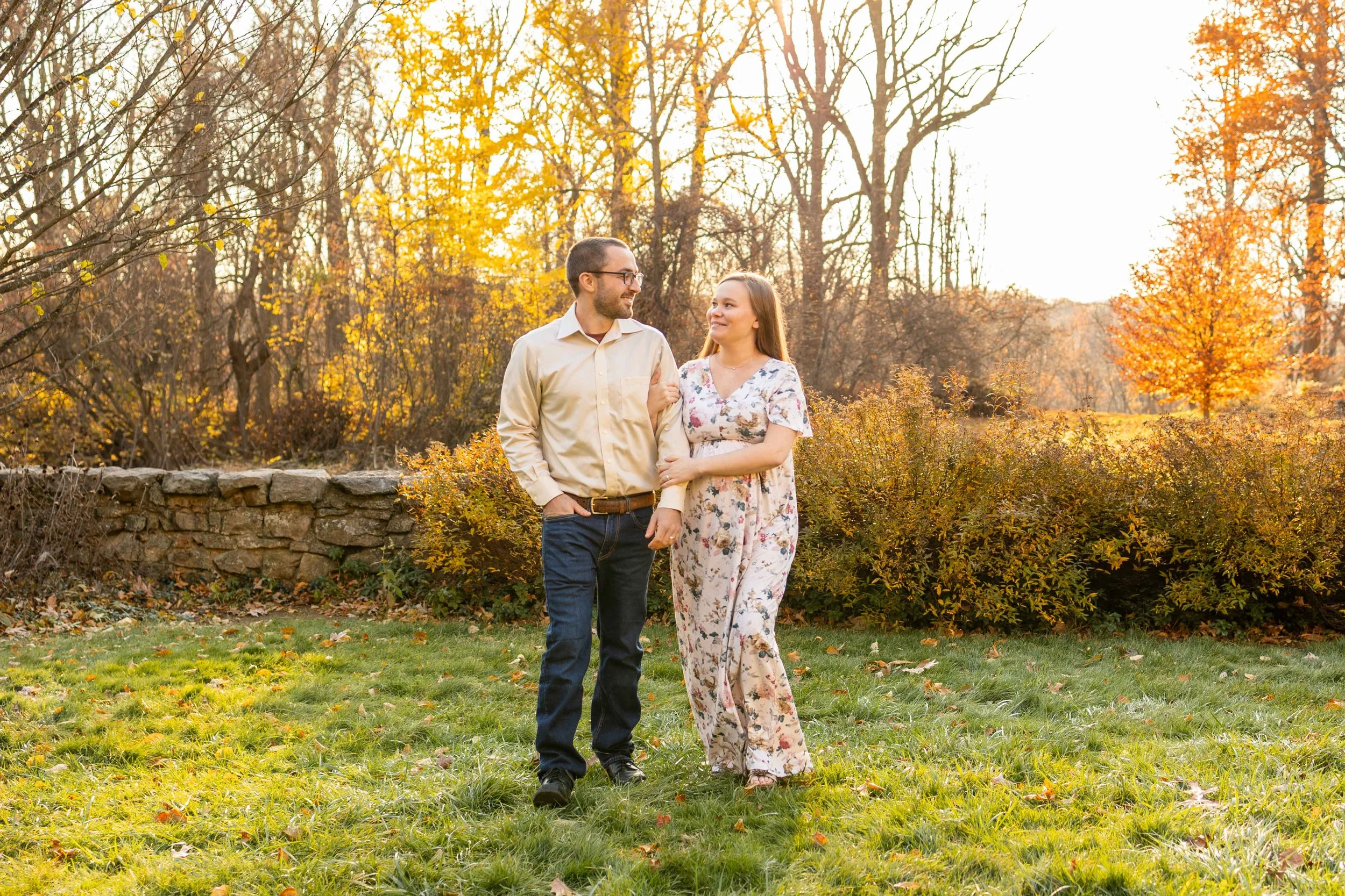 An expecting couple wearing a floral dress, white shirt, and jeans walks together outdoors at golden hour - photo taken by Jennifer Fulton Photography in Central Jersey