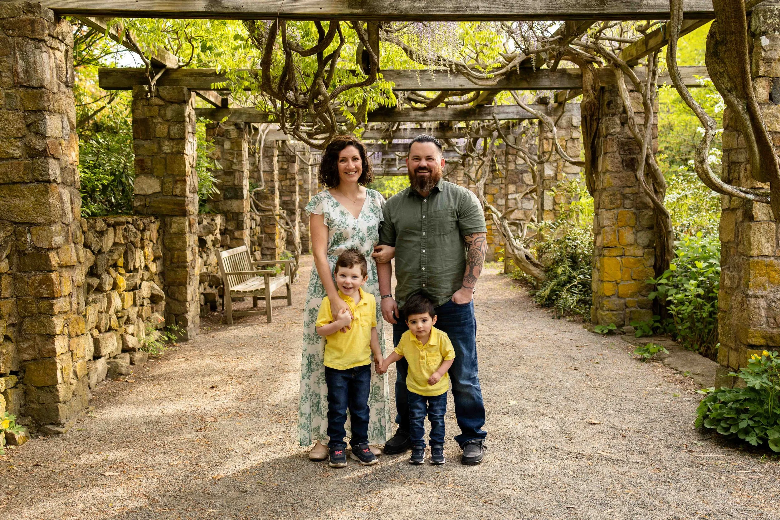 A family of four stands under a wisteria topped pergola - family photo taken by Jennifer Fulton Photography in Somerset County NJ