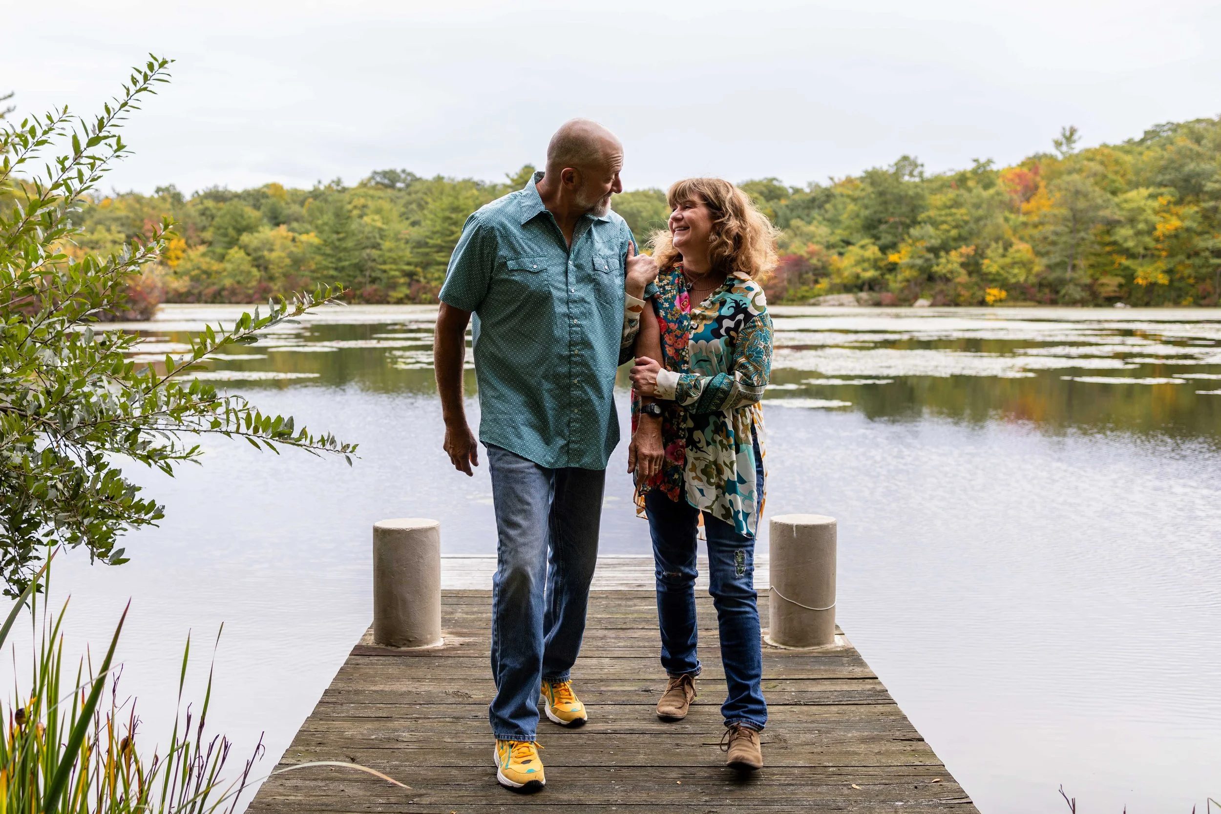 A couple wearing teal, yellow, pink, blue, and white walk on a dock at a lake - couples photo taken by Jennifer Fulton Photography in Mercer County