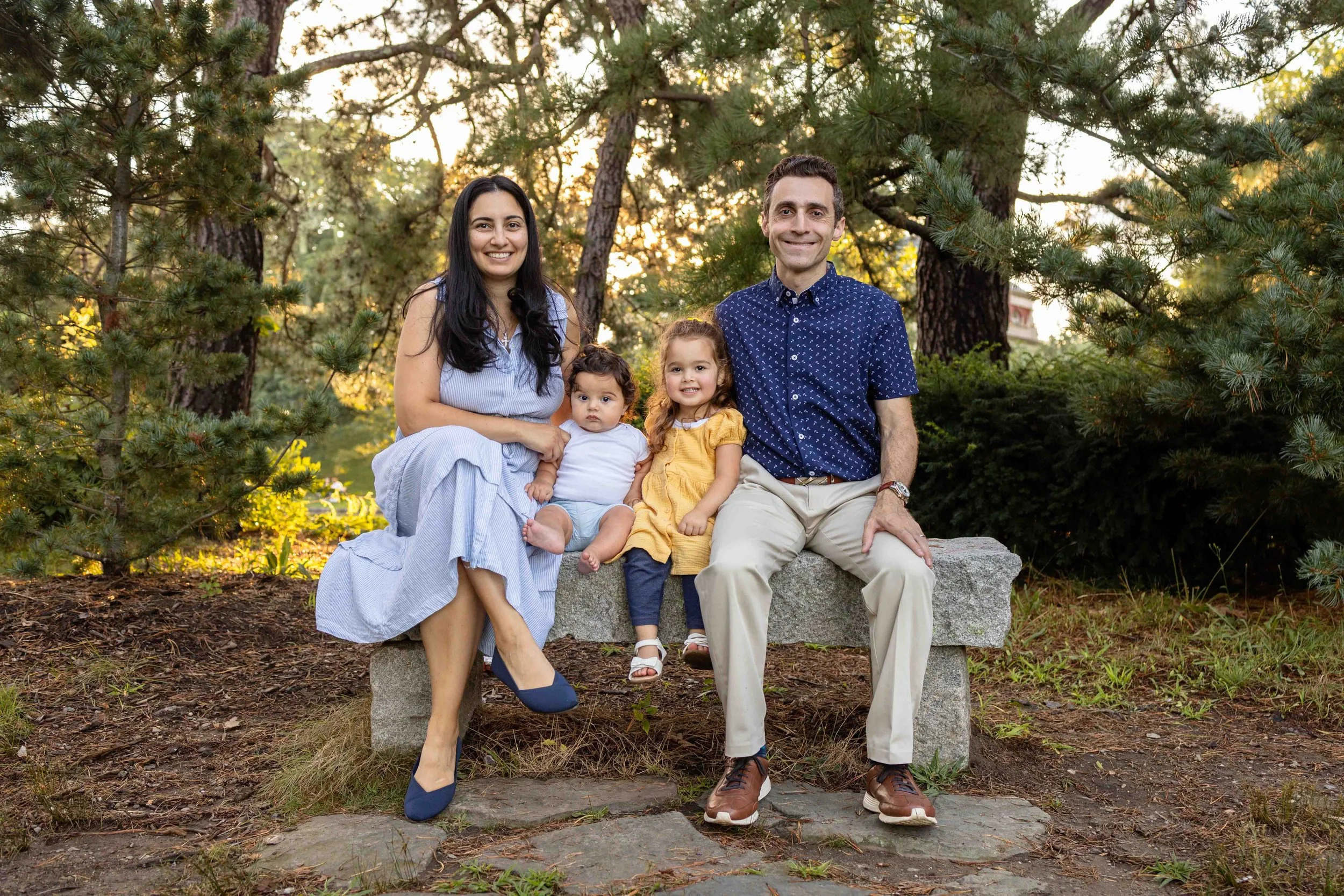 A family of four sits on a bench outdoors at golden hour while wearing blue, yellow, and white outfits - family photo taken by Jennifer Fulton Photography in Central New Jersey