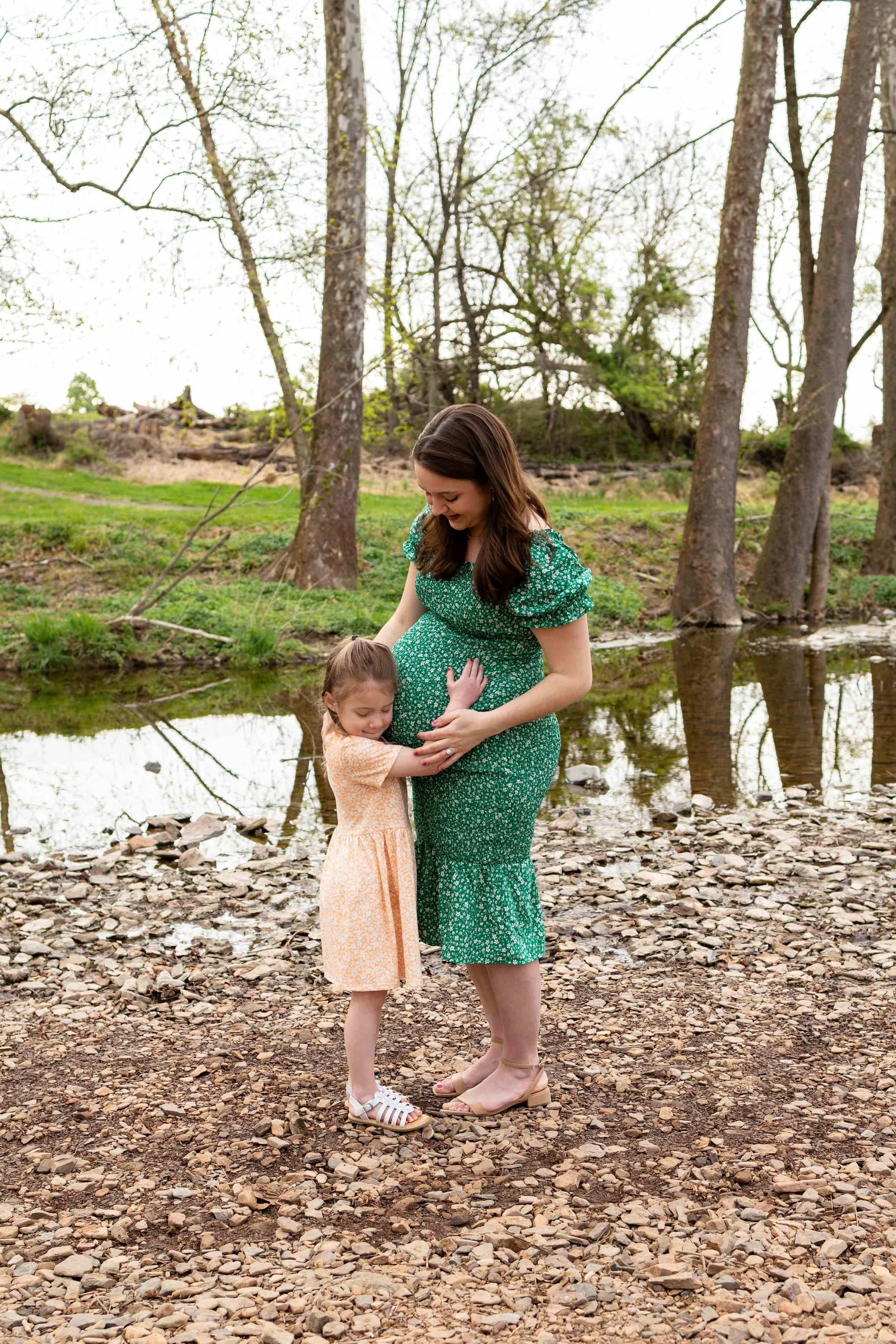 Expecting mom and her daughter hug while standing by a creek - maternity photo taken by Jennifer Fulton Photography in Flemington, NJ