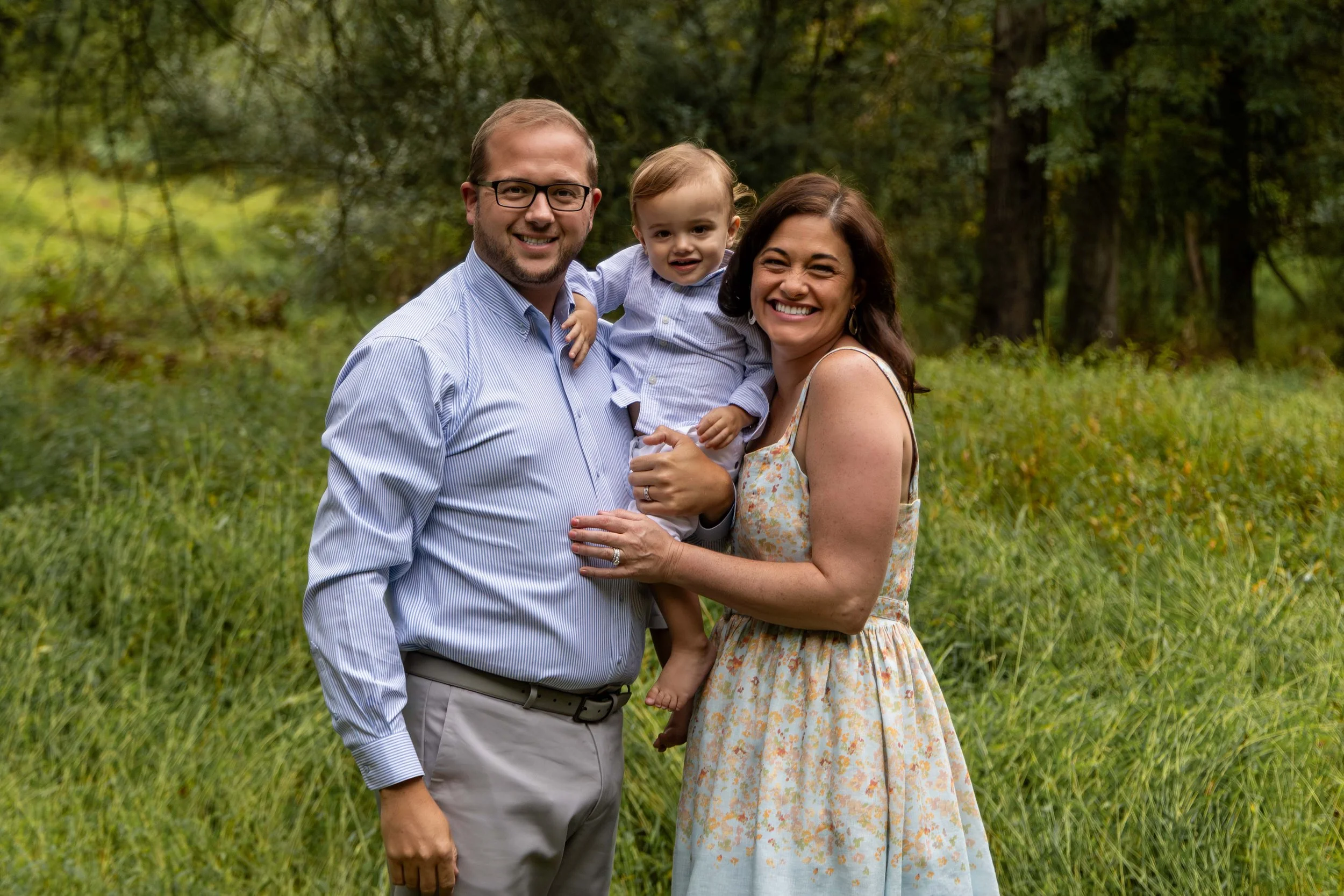 A young family of three stand in a field wearing blue, gray, and floral outfits. Family photo taken by Jennifer Fulton Photography in Hunterdon County, New Jerey